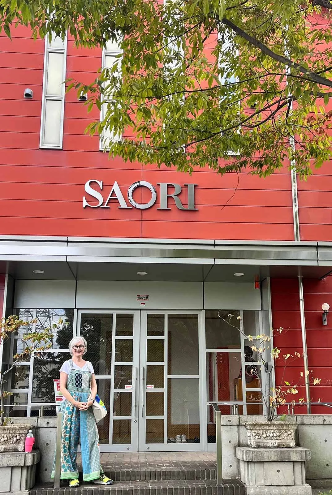A woman wearing glasses and a colorful dress standing in front of the Saori building entrance, which has a red facade with mail slots and a glass door, tree branches overhead, and concrete planters on either side.