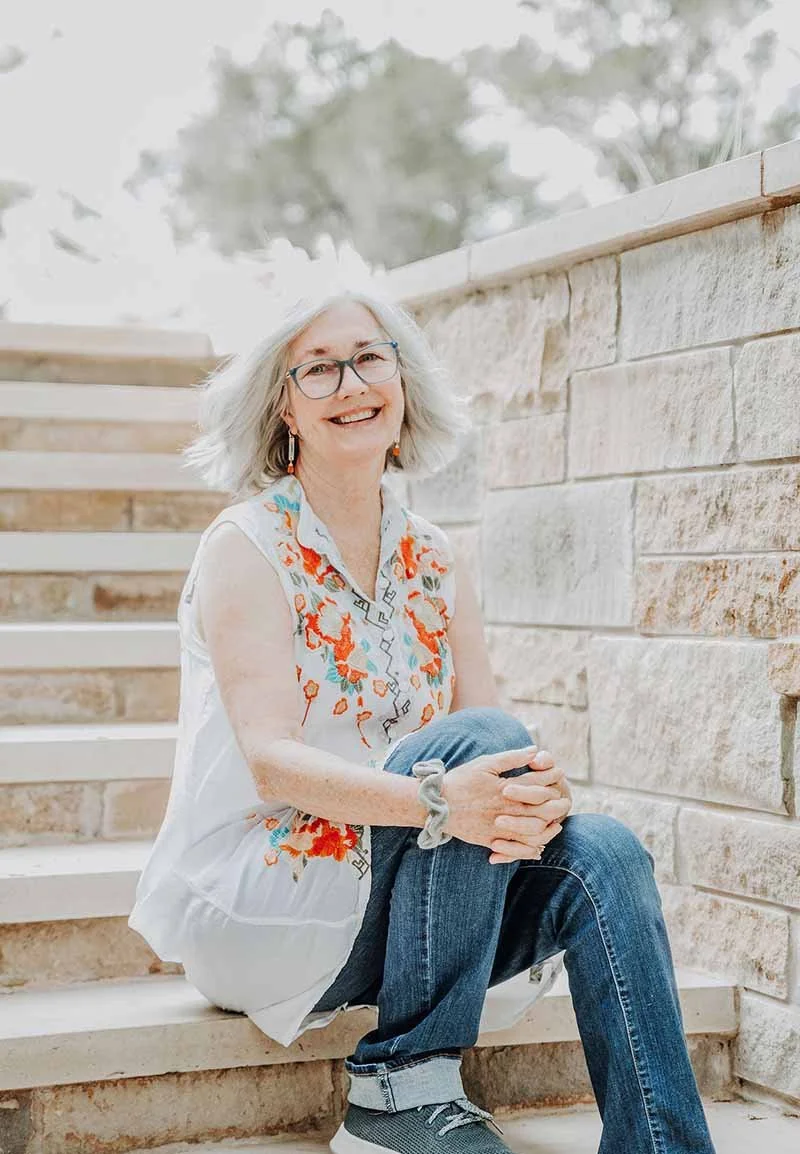 An older woman with gray hair, glasses, and earrings sitting on outdoor stairs next to a stone wall, smiling, wearing a white top with colorful floral embroidery and blue jeans.