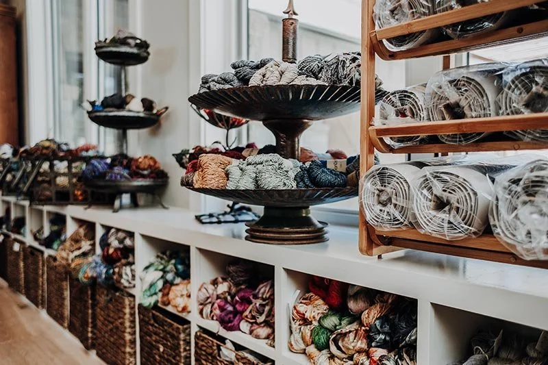 Display of various yarn balls organized on a white shelf with baskets and decorative stands.