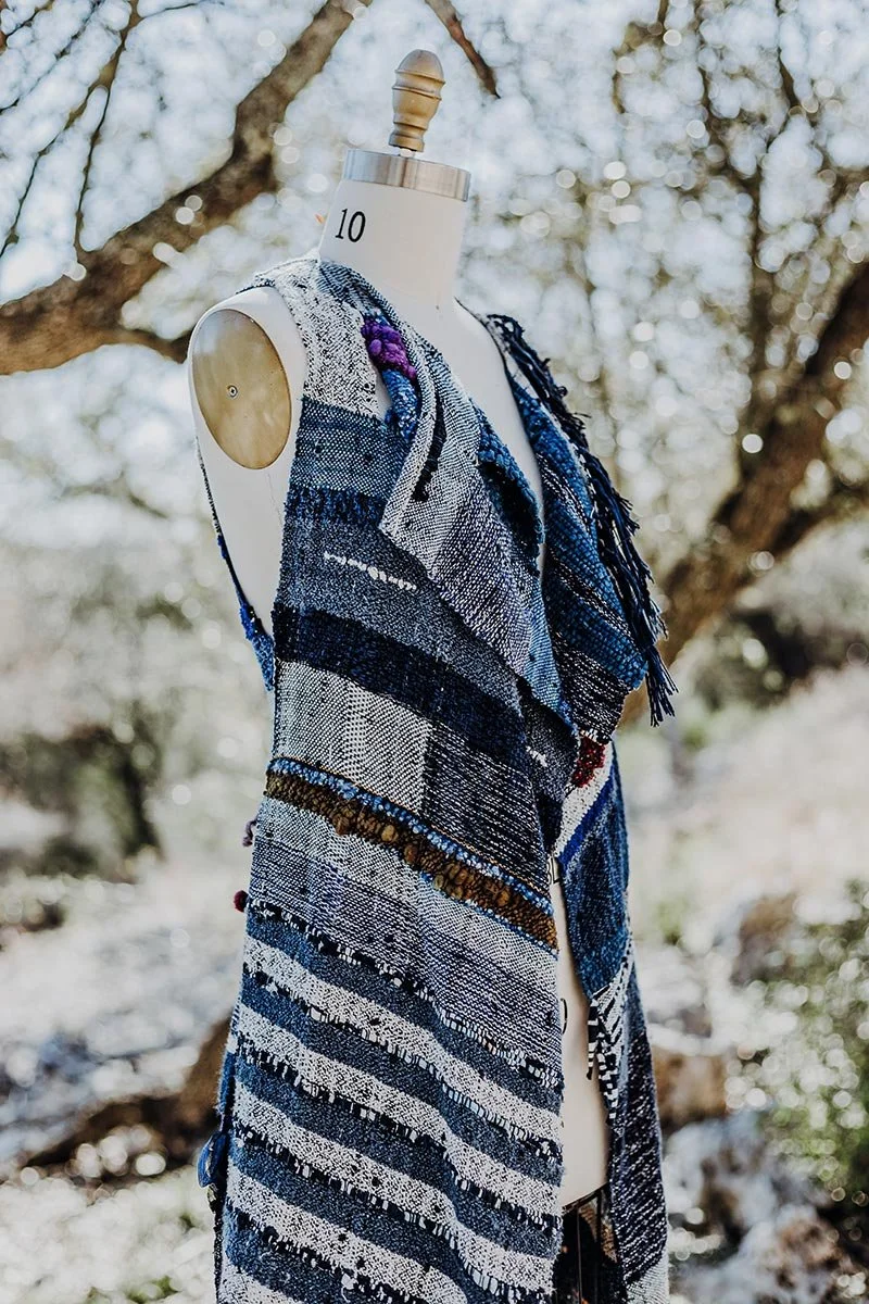 A dress made of various shades of blue, white, and black woven fabric displayed on a mannequin outdoors with snow and tree branches in the background.