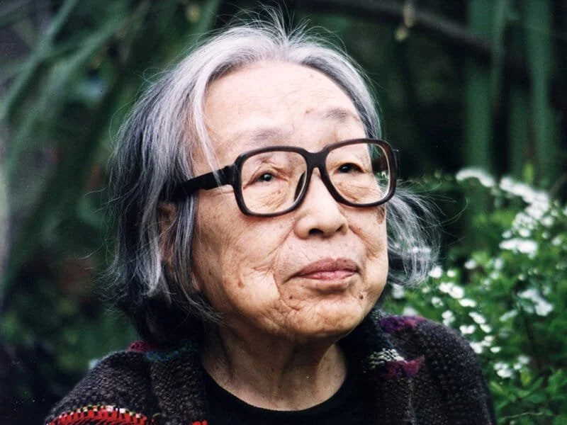 Close-up of an elderly woman with gray hair, wearing glasses, outdoors in a garden.