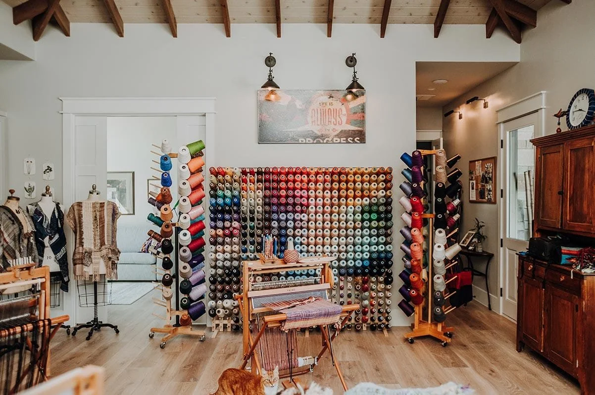 Interior of a craft room with a wall of colorful yarn spools on wooden racks, a small weaving loom with yarn, dress forms with knitted garments, and other crafting supplies.