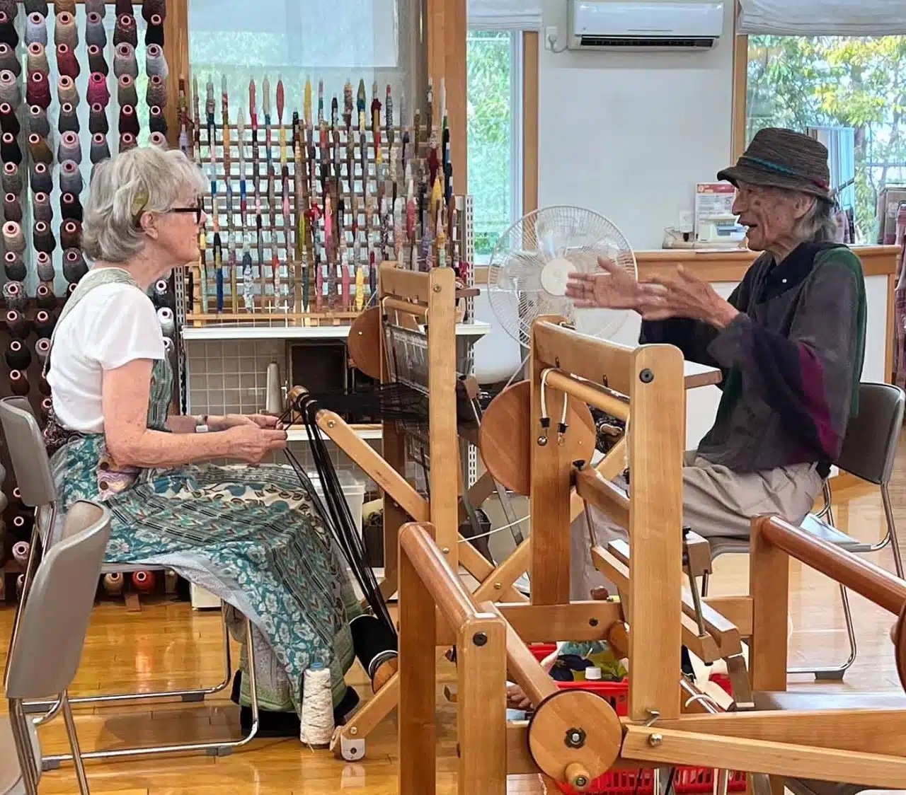 Two elderly women are sitting across from each other in a craft or sewing room, with one woman actively demonstrating or explaining something while the other listens attentively. They are separated by a wooden frame or loom, and there are various spools of thread and craft supplies in the background, with a fan and large window visible.
