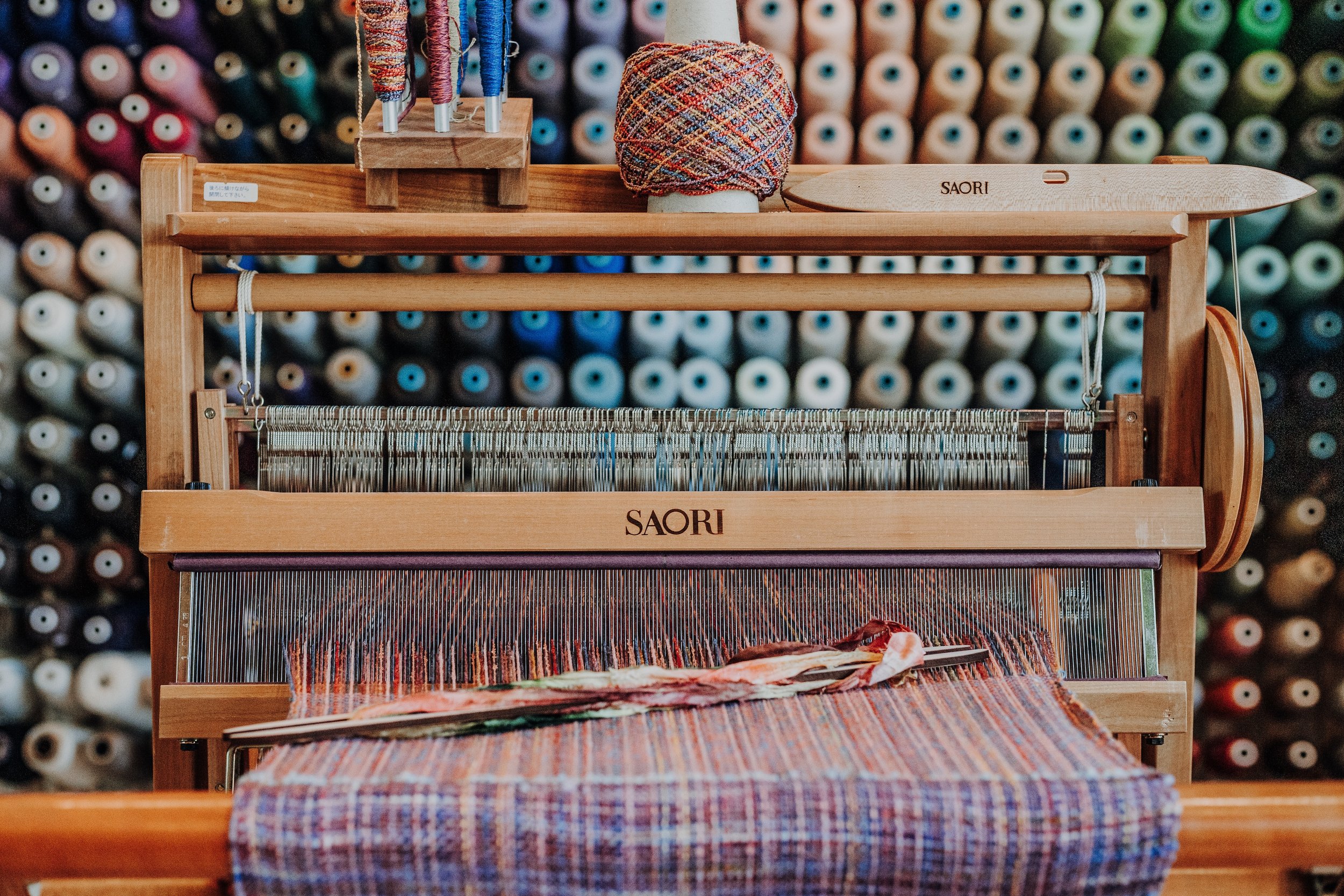 A wooden loom with colorful thread, displayed in front of a wall of multicolored thread spools.