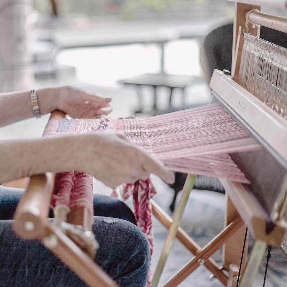 A person weaving on a small loom, working with pink and red threads. The loom is set up on a wooden table in a bright, indoor space.