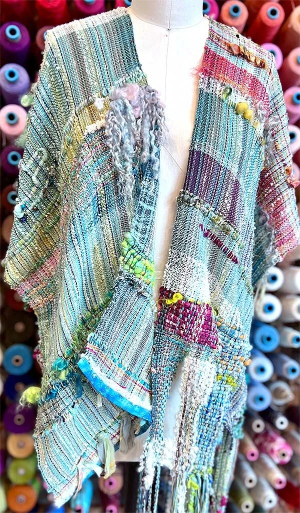 Colorful, patchwork-style woven garment displayed on a mannequin, with spools of thread in various colors in the background.