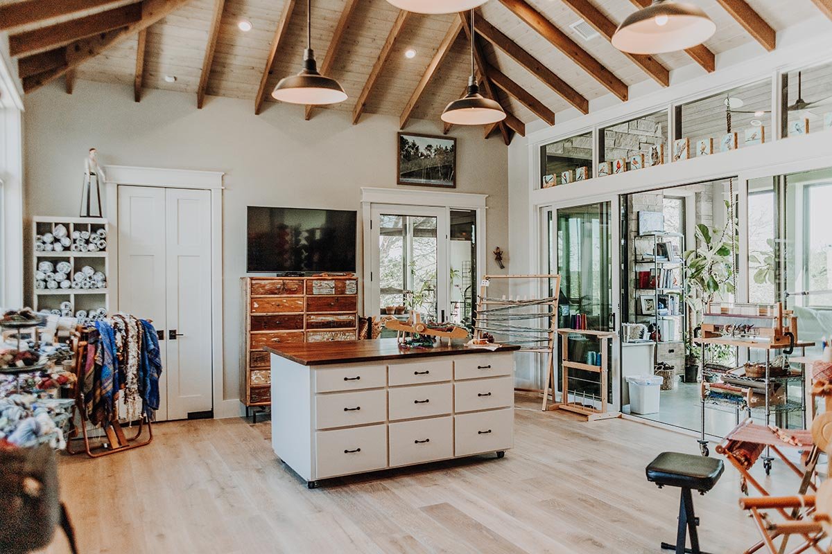 A craft room featuring a high wooden ceiling with exposed beams, a large window wall with sliding doors, and various craft supplies including yarn, fabric, and spinning wheels.