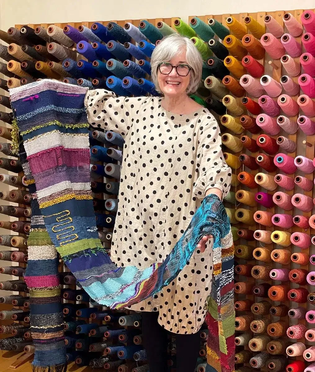 A woman with short gray hair and glasses smiling, standing in front of a wall of colorful thread spools, holding a multicolored knitted or woven scarf with her right arm extended.
