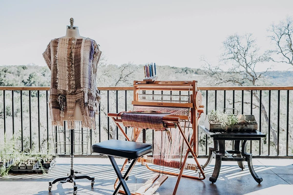 A balcony with a mannequin dressed in a fringed woven top or shawl, a weaving loom with colorful threads, a black table with plants and gardening tools, and a view of trees and sky in the background.