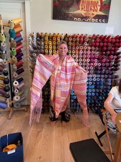 Person standing in front of a wall of colorful spools of thread in a sewing or craft room, holding a large, pink, and yellow striped shawl or scarf.