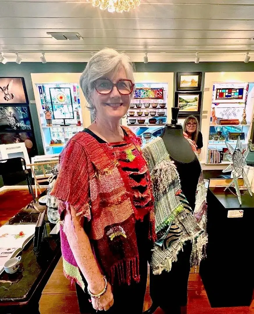 Woman with grey hair and red glasses wearing a colorful, textured, patchwork-style top, standing inside an art or craft store with various decorative items and artworks in the background.