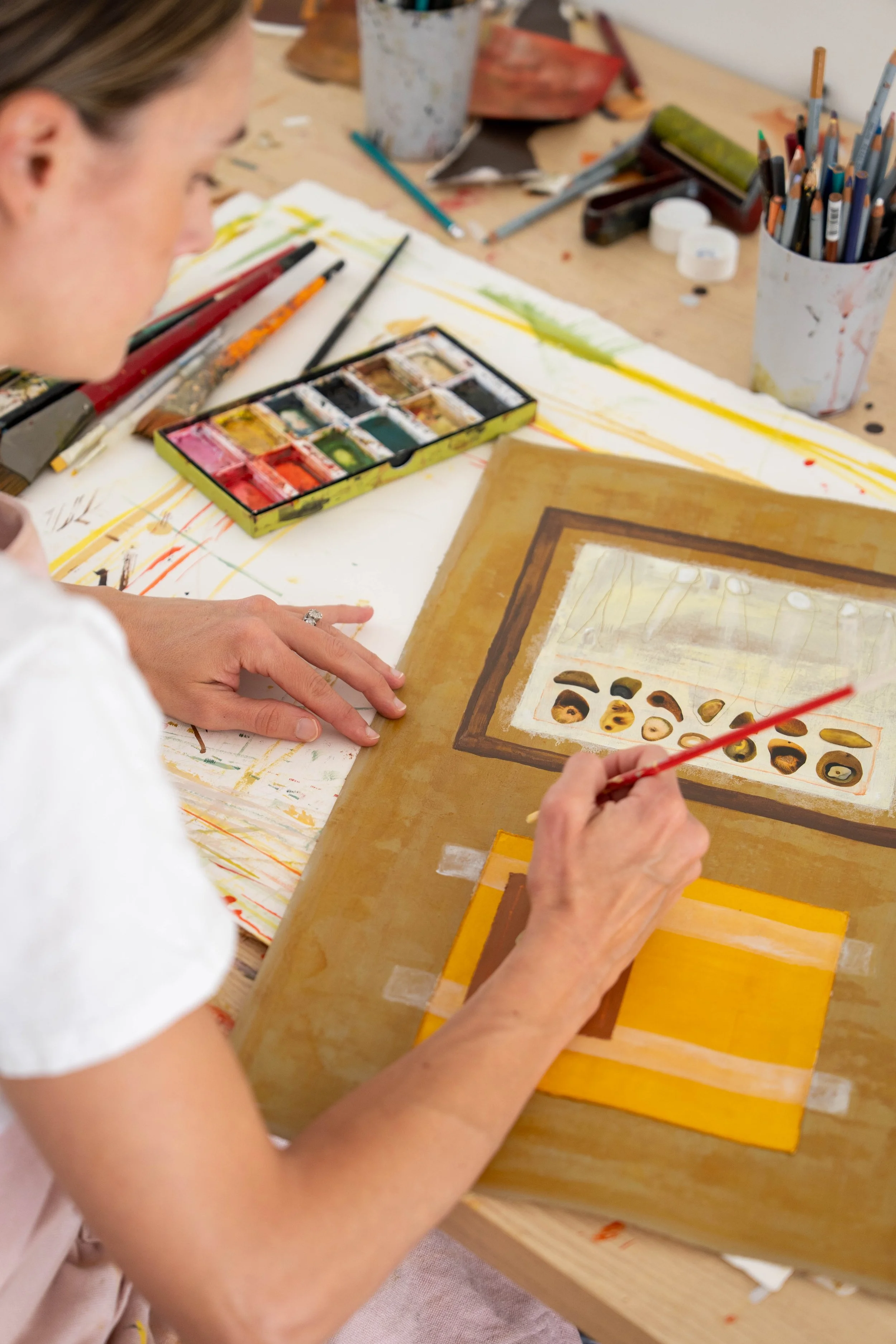 Woman painting a picture with watercolors on paper, surrounded by art supplies on a wooden table.