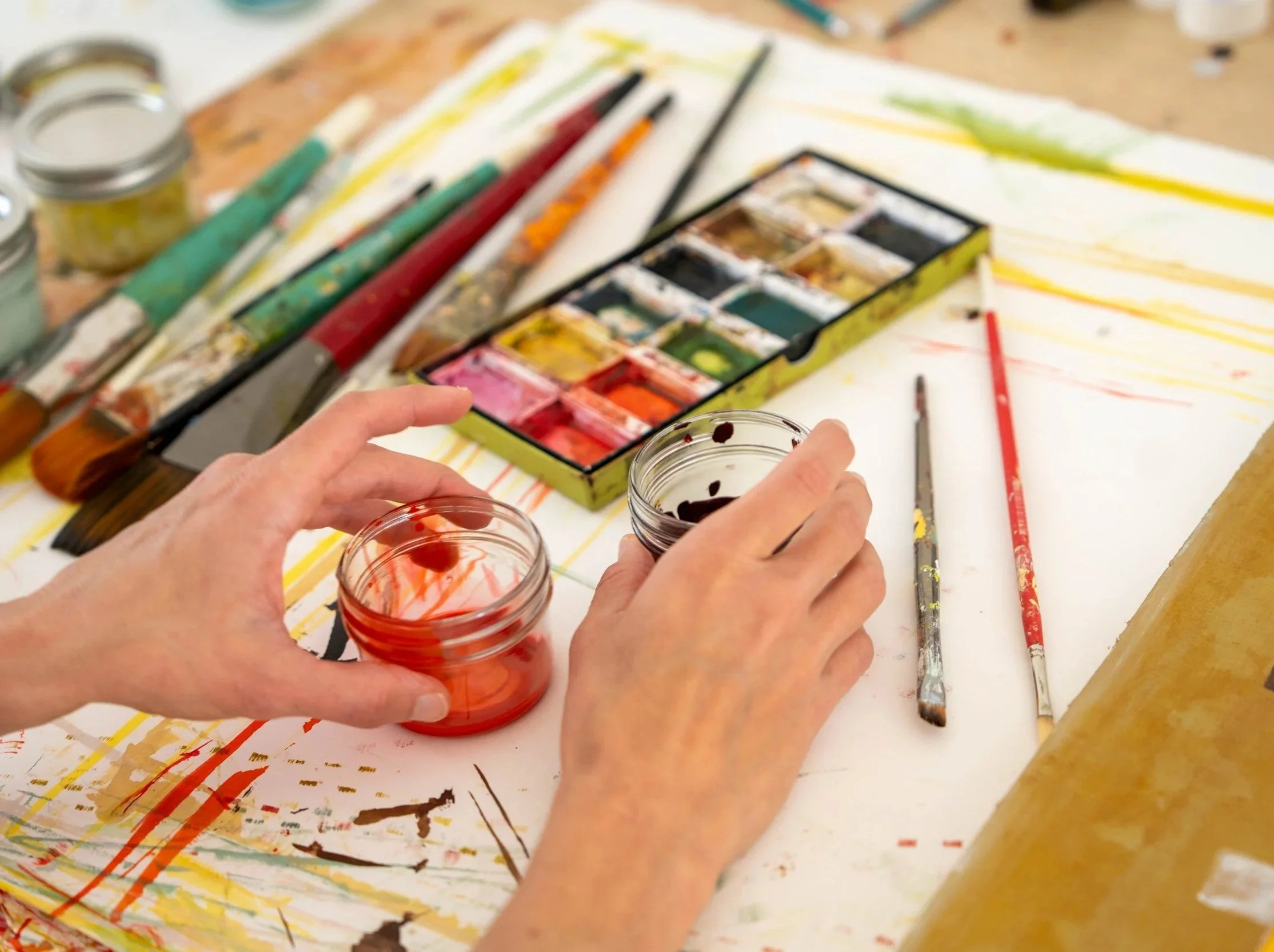 Person holding small jar with red paint in an art studio with watercolor paints and paintbrushes on a colorful painted table.