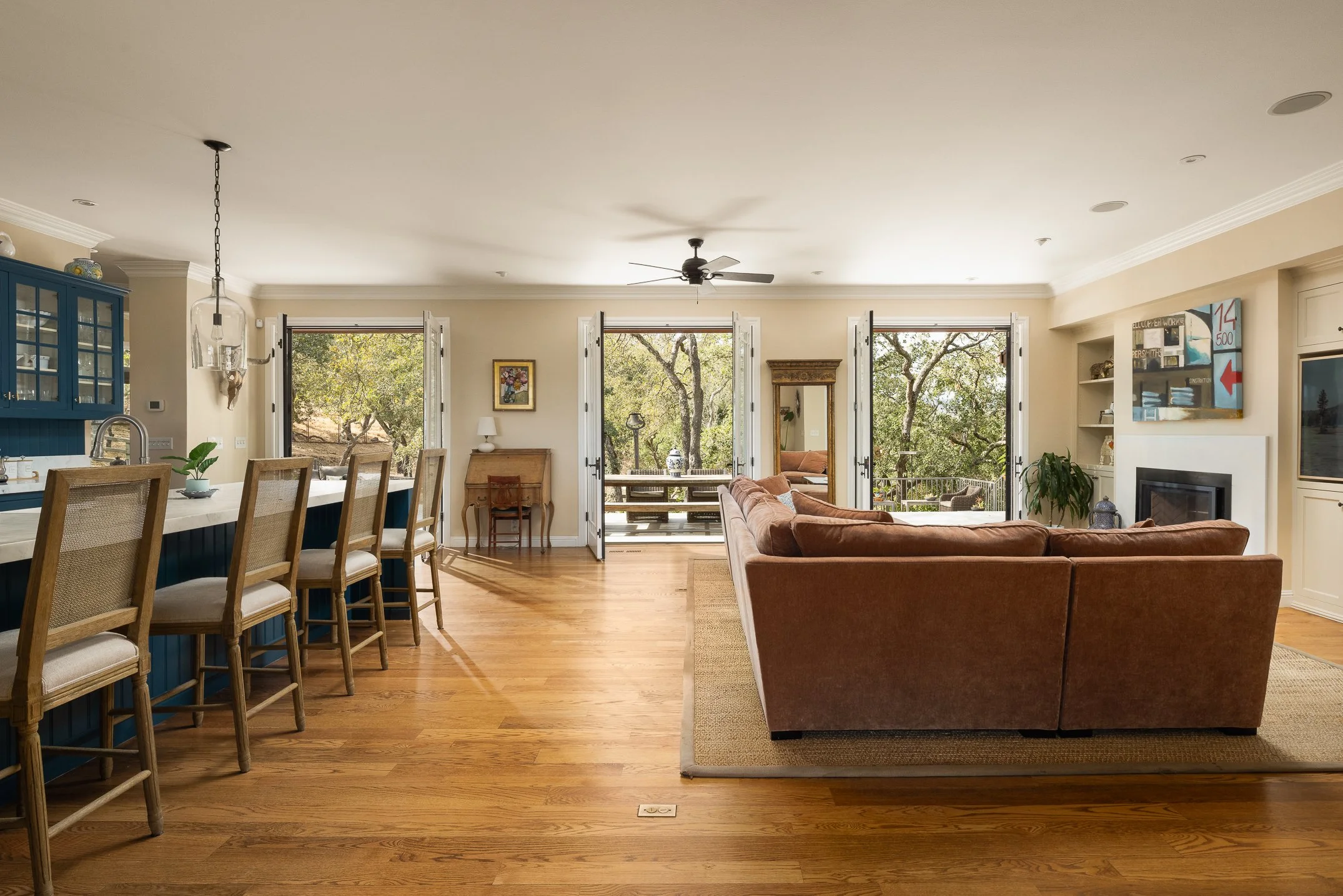 Spacious living room with open doors to balcony, featuring a large brown couch, modern artwork, a ceiling fan, and hardwood floors.
