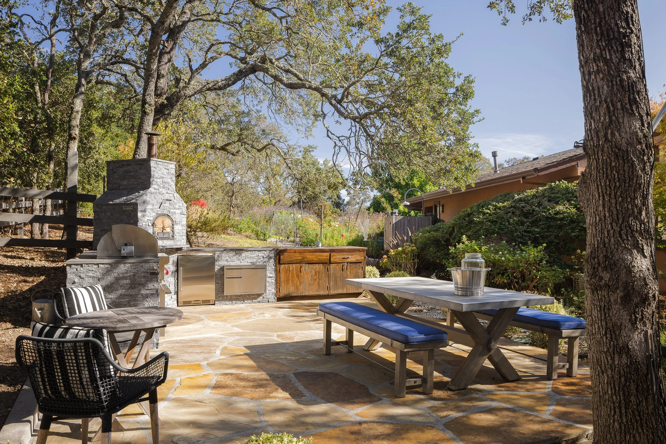 A backyard patio area with outdoor dining furniture, a stone outdoor grill or fireplace, and surrounding trees and plants under a blue sky.