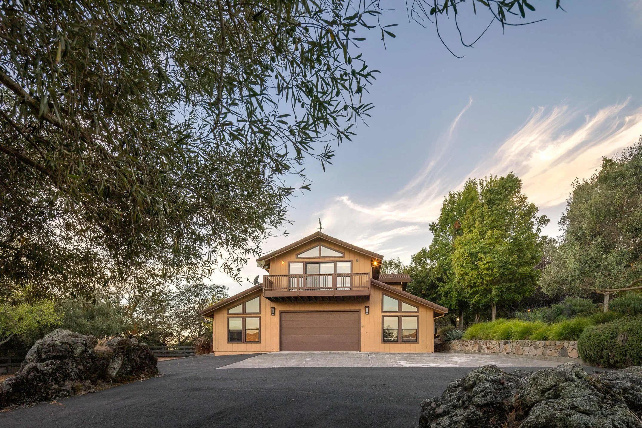 A two-story house with a brown garage door, surrounded by trees and rocks, under a partly cloudy sky during evening or early morning.