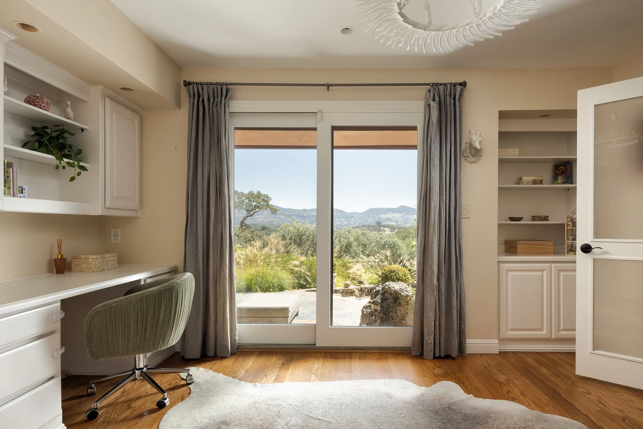 A room with sliding glass doors opening to a scenic outdoor view with hills and trees. The room features built-in white shelves, gray curtains, a desk area, a green swivel chair, a ceiling light fixture, and a beige carpet on wooden flooring.