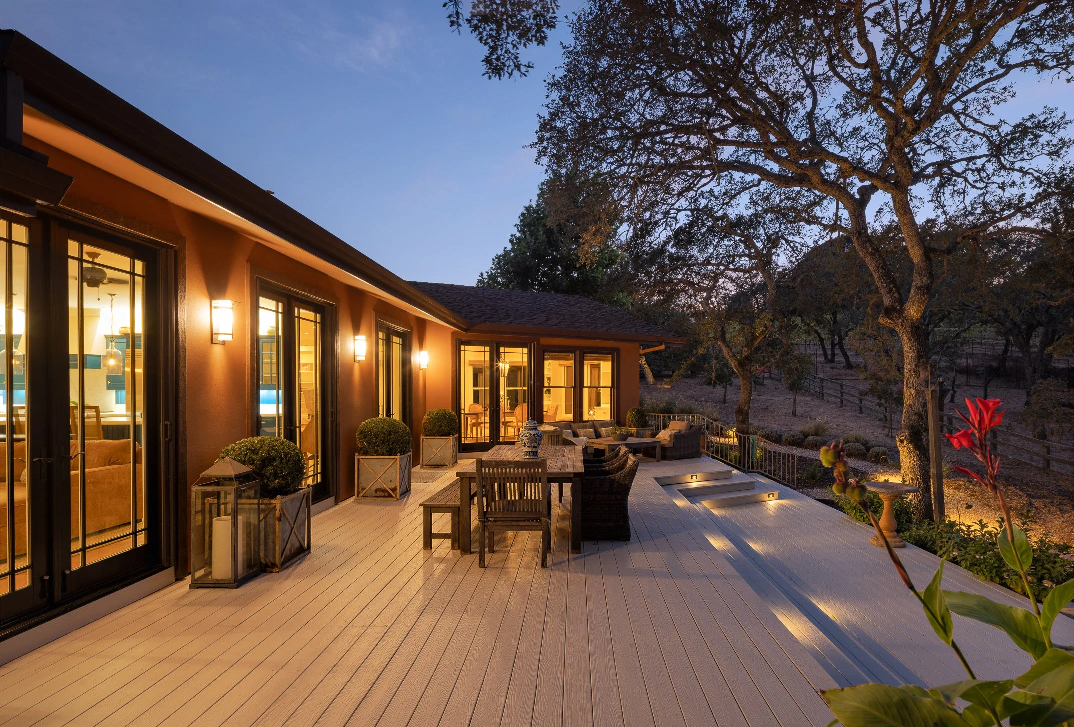 Residential house's outdoor patio area at dusk, with large glass doors, warm porch lighting, outdoor furniture, and a large tree in the background.