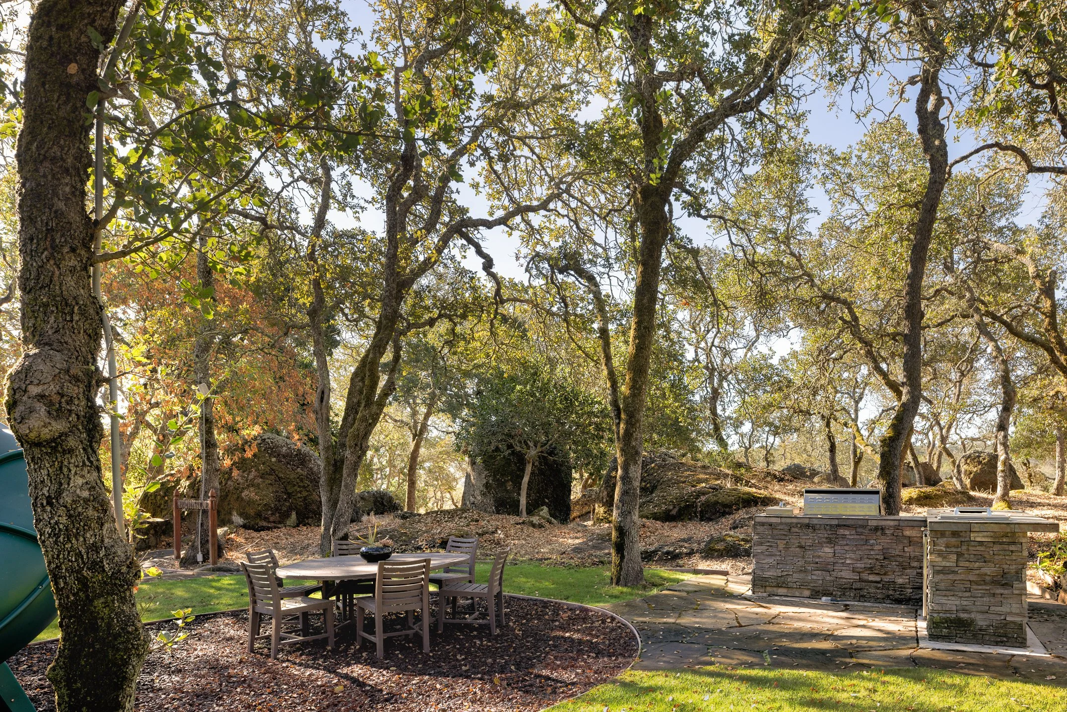 Backyard with large trees, a round dining table with chairs, a stone outdoor kitchen, and rocky landscaping under a clear sky.