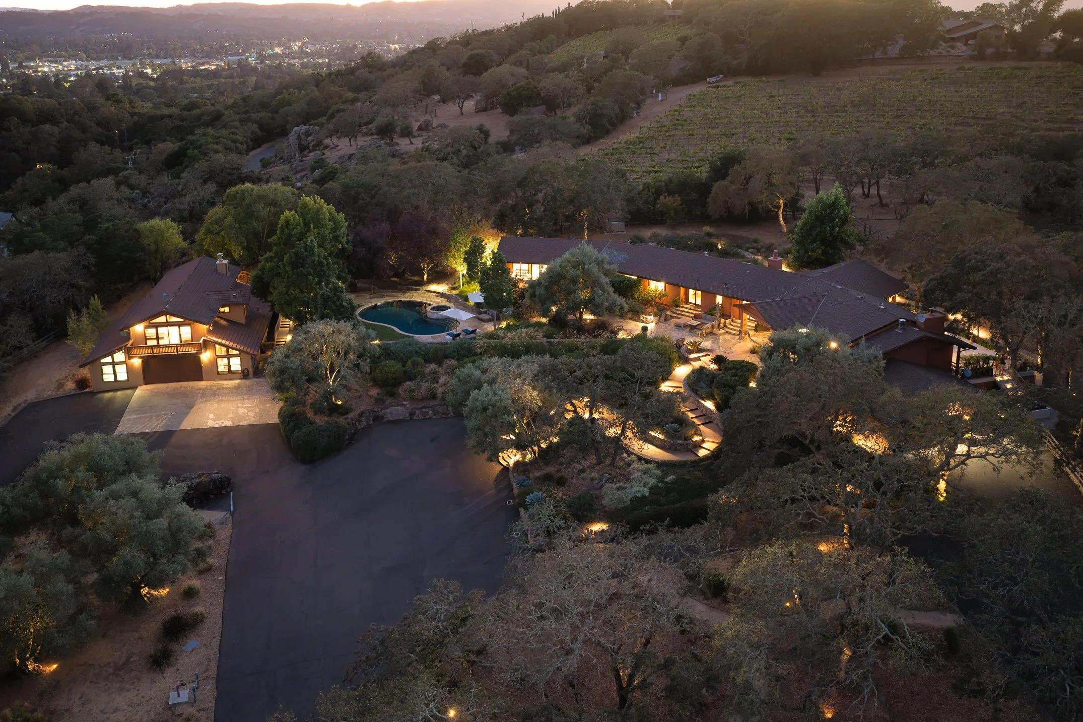 An aerial view of a residential area at dusk showing houses with lit windows, a swimming pool, surrounding trees, and hills in the background.