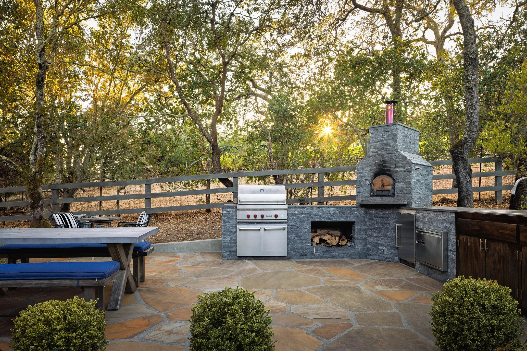 An outdoor patio area with a wood-fired oven, barbecue grill, and outdoor kitchen setup, surrounded by trees with sunlight peeking through.