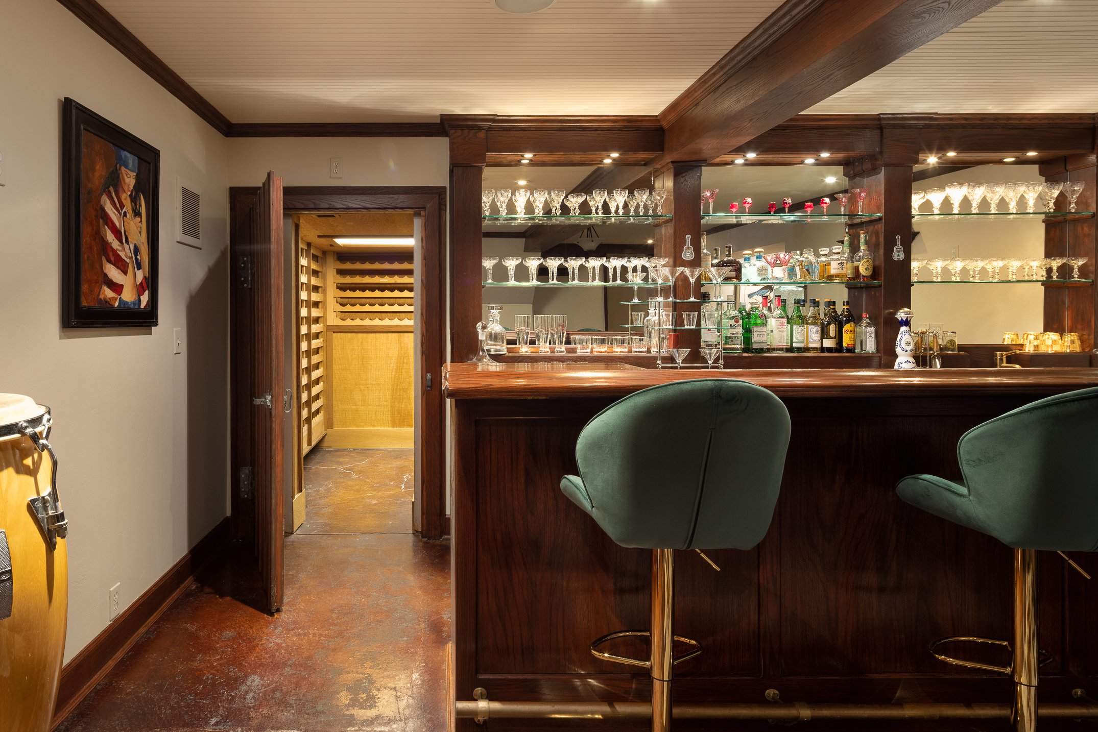 Image of a home bar area with glass shelves holding various liquor bottles and glassware, and green velvet bar stools in the foreground. Opens to wine cellar.