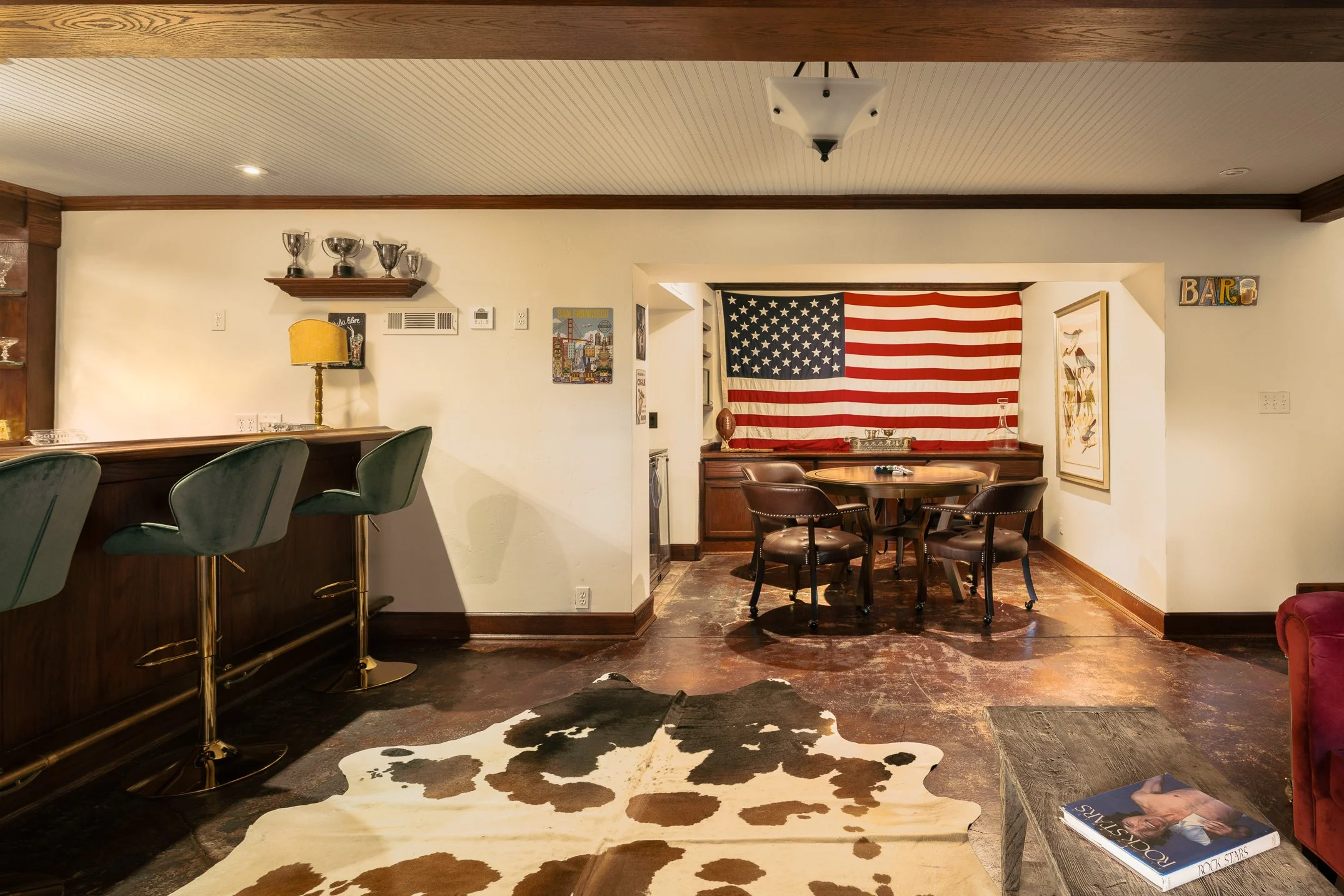 A cozy dining area with a cowhide rug on dark hardwood floor, a round wooden table with black chairs, a wall decorated with American flag, and various wall art and trophies.