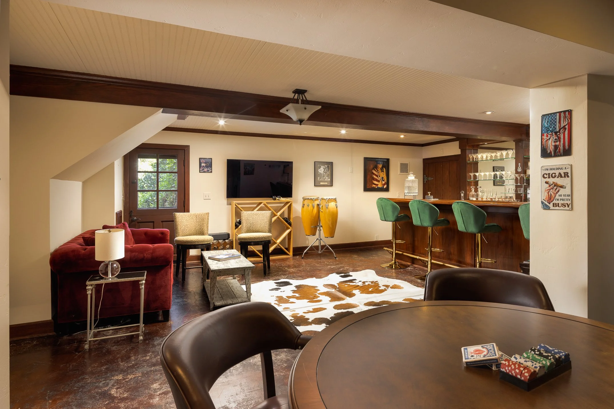 Interior view of a cozy living room with a bar area, featuring green bar stools, artwork on the walls, a television, and musical instruments including bongos. There's a cowhide rug on the wooden floor and a window letting in natural light.