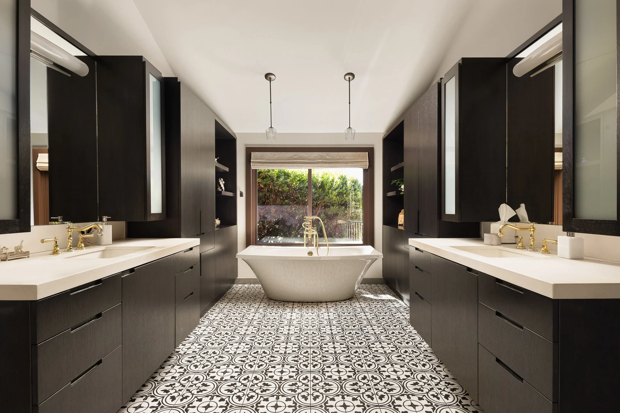 A modern bathroom with black cabinets on both sides, white countertops, a freestanding bathtub, and a large window overlooking greenery, with patterned floor tiles.