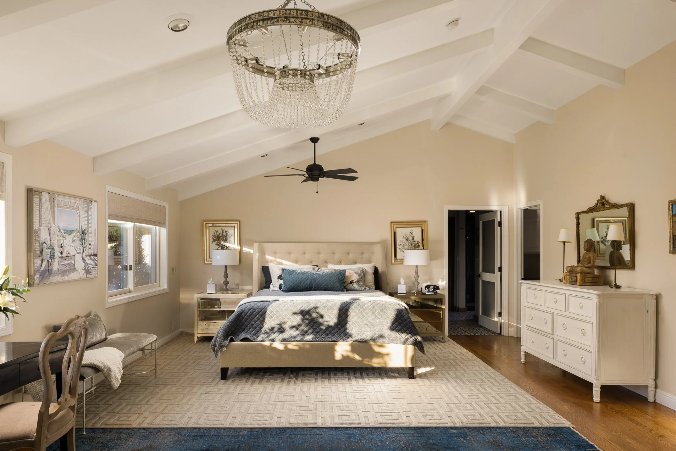 A bedroom with a beige upholstered bed, white nightstands, and lamps. The ceiling has a large crystal chandelier and a black ceiling fan. The room has beige walls, large windows with blinds, and framed artwork. There is a white dresser with decorativ