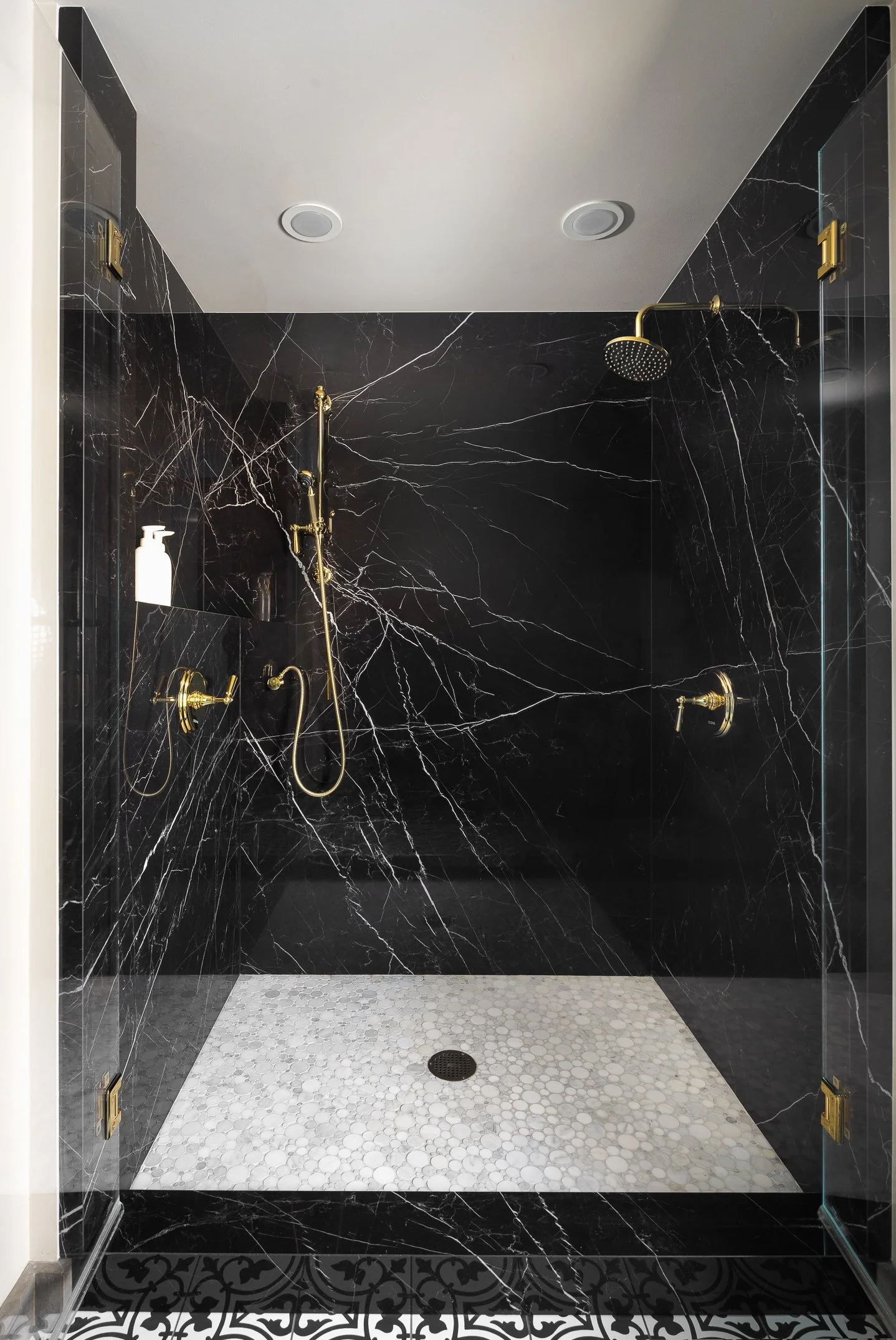 A modern shower with black marble walls, gold fixtures, a handheld showerhead, an overhead rainfall showerhead, and a white pebble tile floor with a black drain.