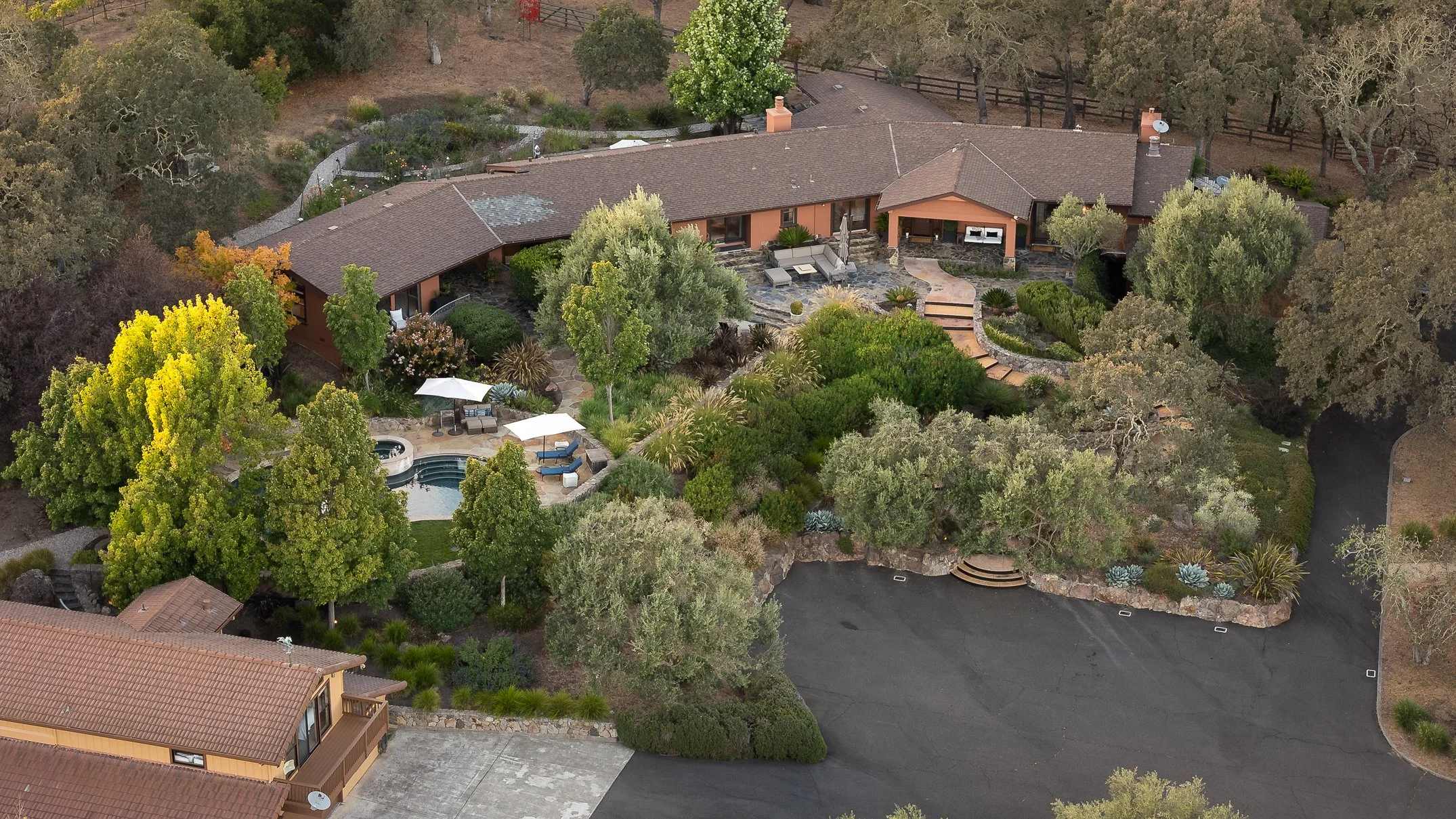 An aerial view of a large, single-story house surrounded by lush greenery and trees, featuring a backyard with a swimming pool, outdoor seating area, and landscaped garden.