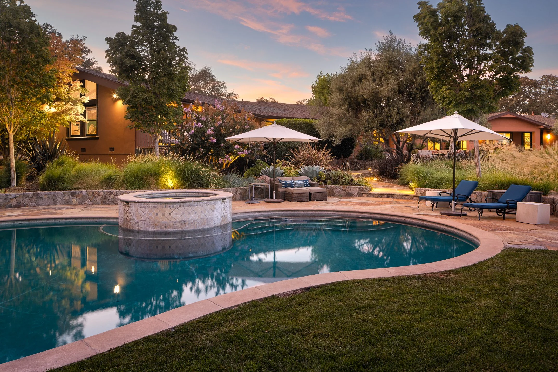 Residential backyard at sunset featuring a swimming pool, poolside furniture, umbrellas, trees, and houses in the background.