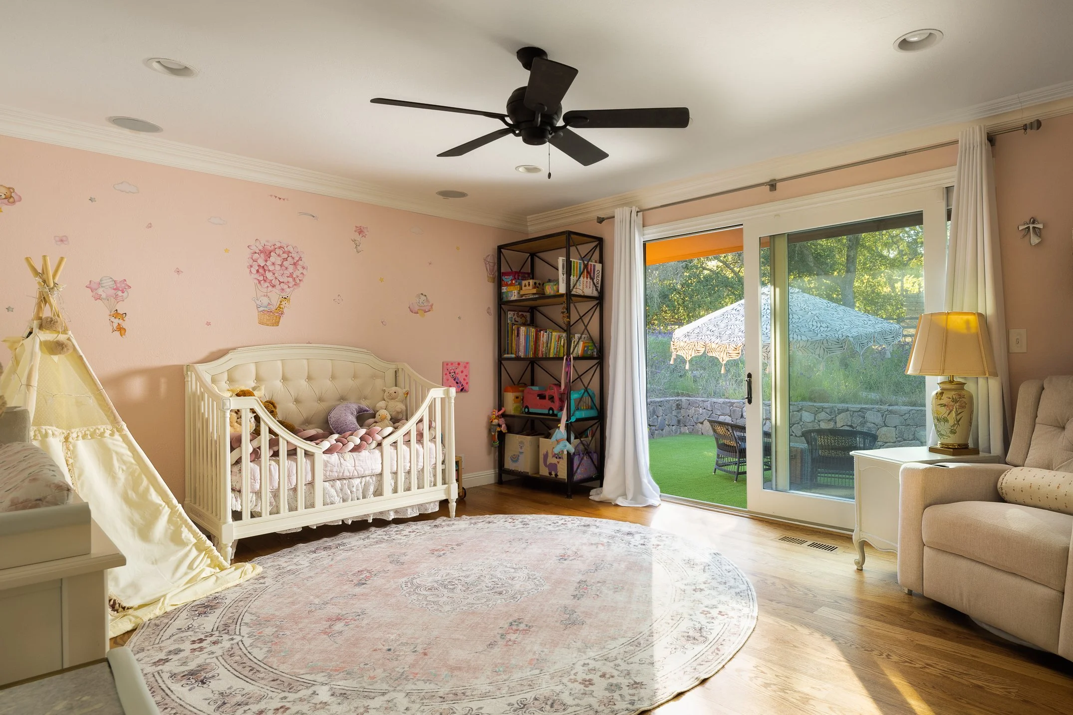 A bright bedroom with pink walls decorated with cute animal and cloud stickers, a white crib filled with stuffed animals, a crib tent, a tall bookshelf, and a large glass sliding door leading to a patio with outdoor furniture and a sun umbrella.