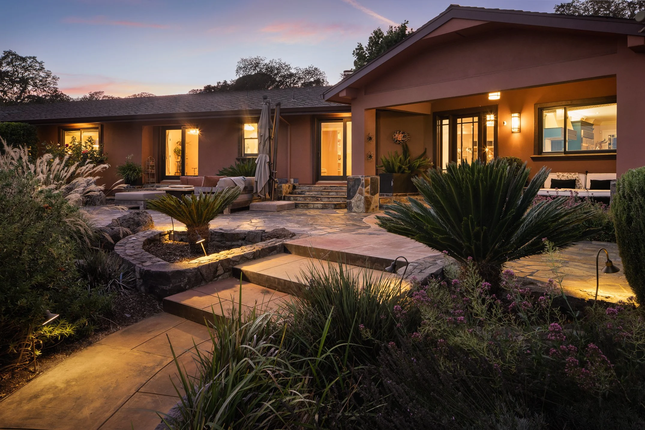 A backyard patio area at dusk with illuminated outdoor lights. The scene includes a stone pathway, lush plants, and an outdoor seating area with cushions. The house has large windows and sliding doors, and the sky shows a sunset with soft pink and pu