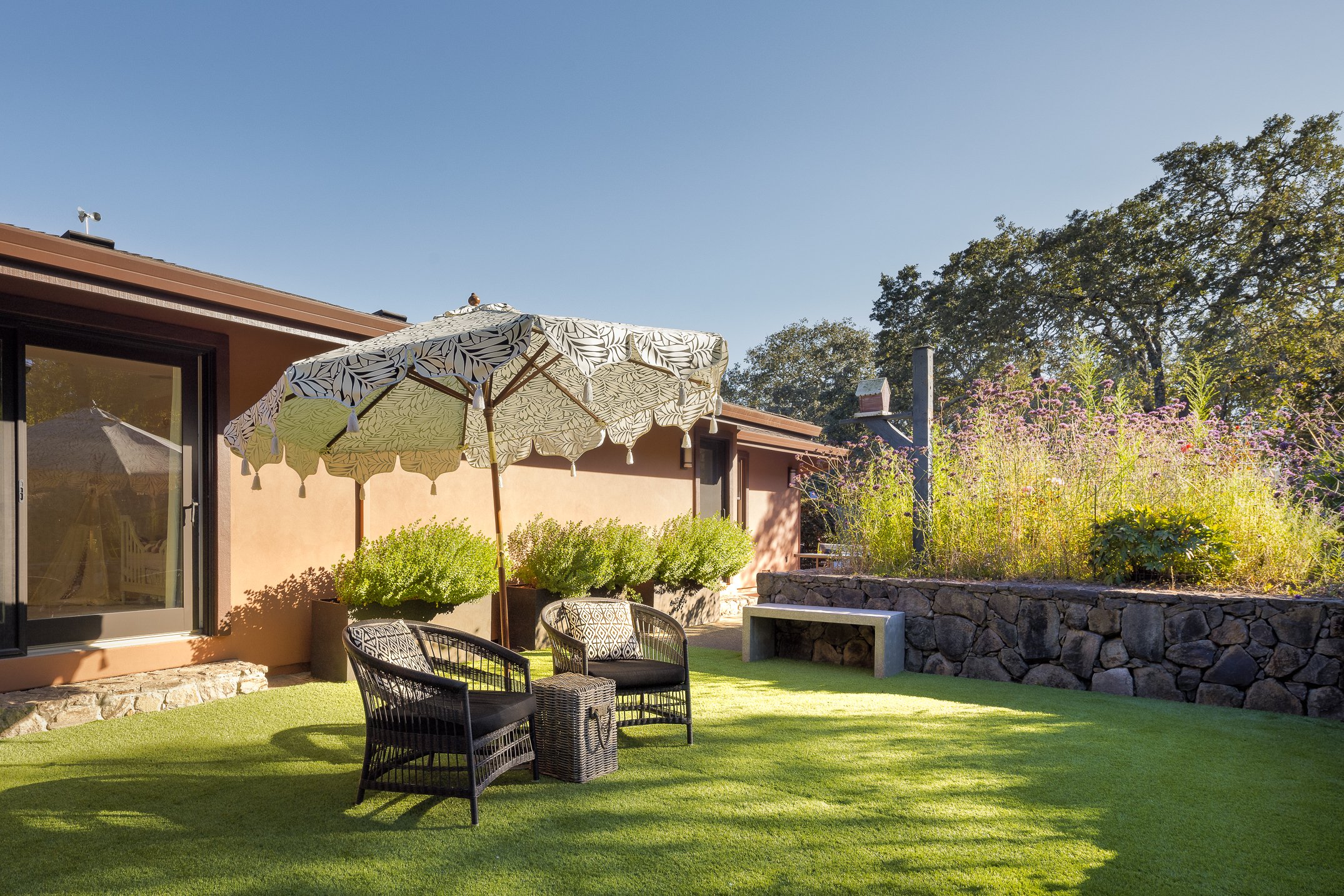 A backyard patio area with outdoor seating, a large umbrella, a pink stucco house, greenery, trees, and flowering plants under a clear blue sky.
