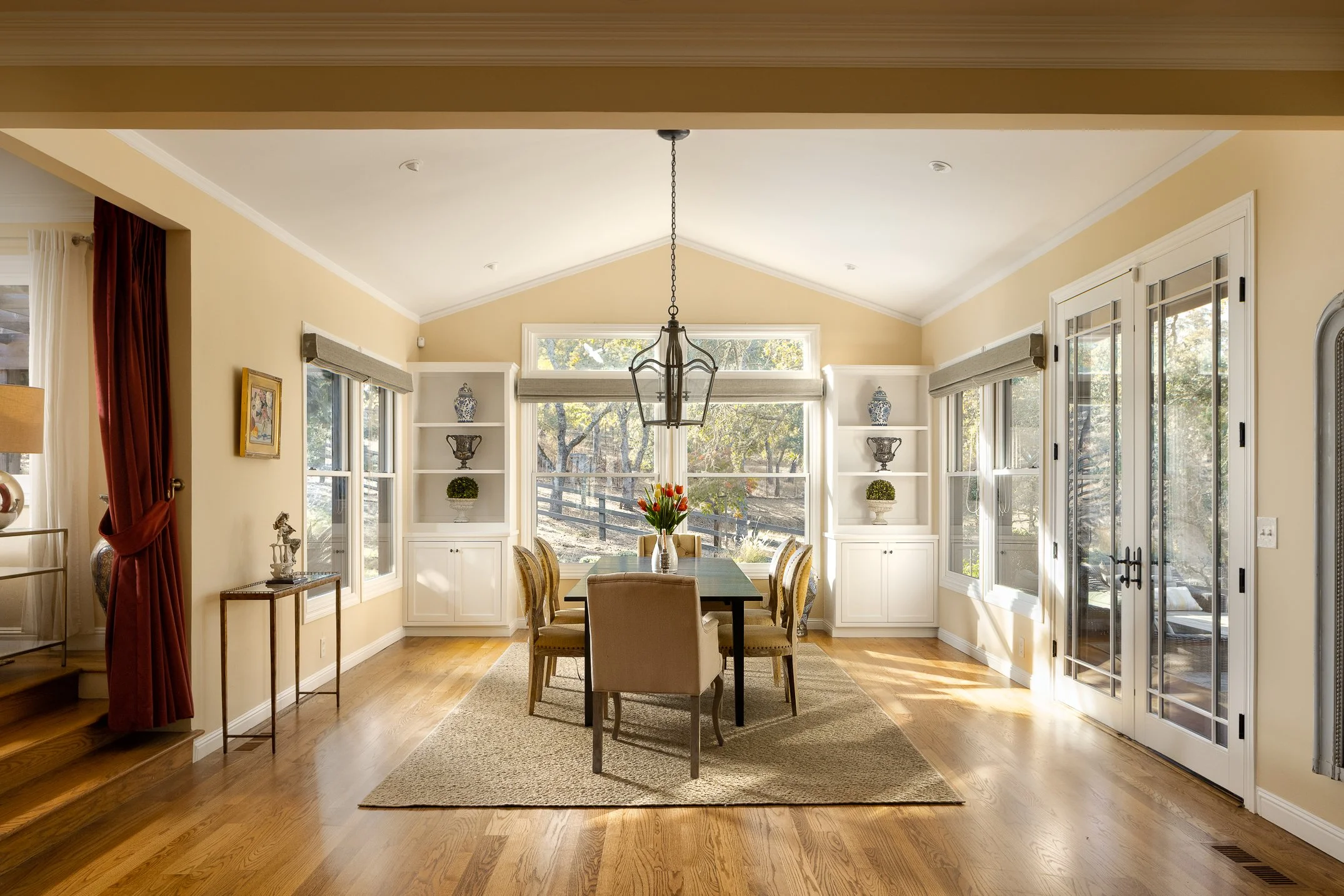 A bright dining room with large windows and a central wooden dining table surrounded by six beige upholstered chairs. A modern metal chandelier hangs above the table. The room features built-in white shelves with decorative items and has hardwood flo