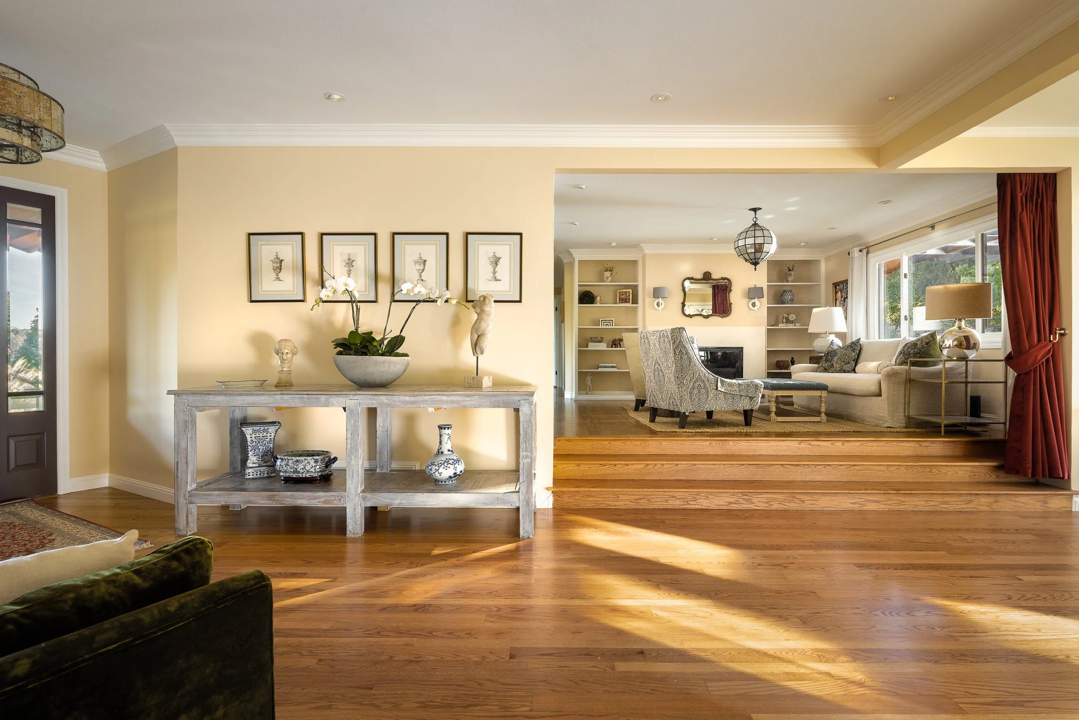 Living room with beige walls, hardwood flooring, and large windows with red curtains. Furniture includes sofa, armchair, lamps, and decorative items, with artwork and decor on shelves and walls.