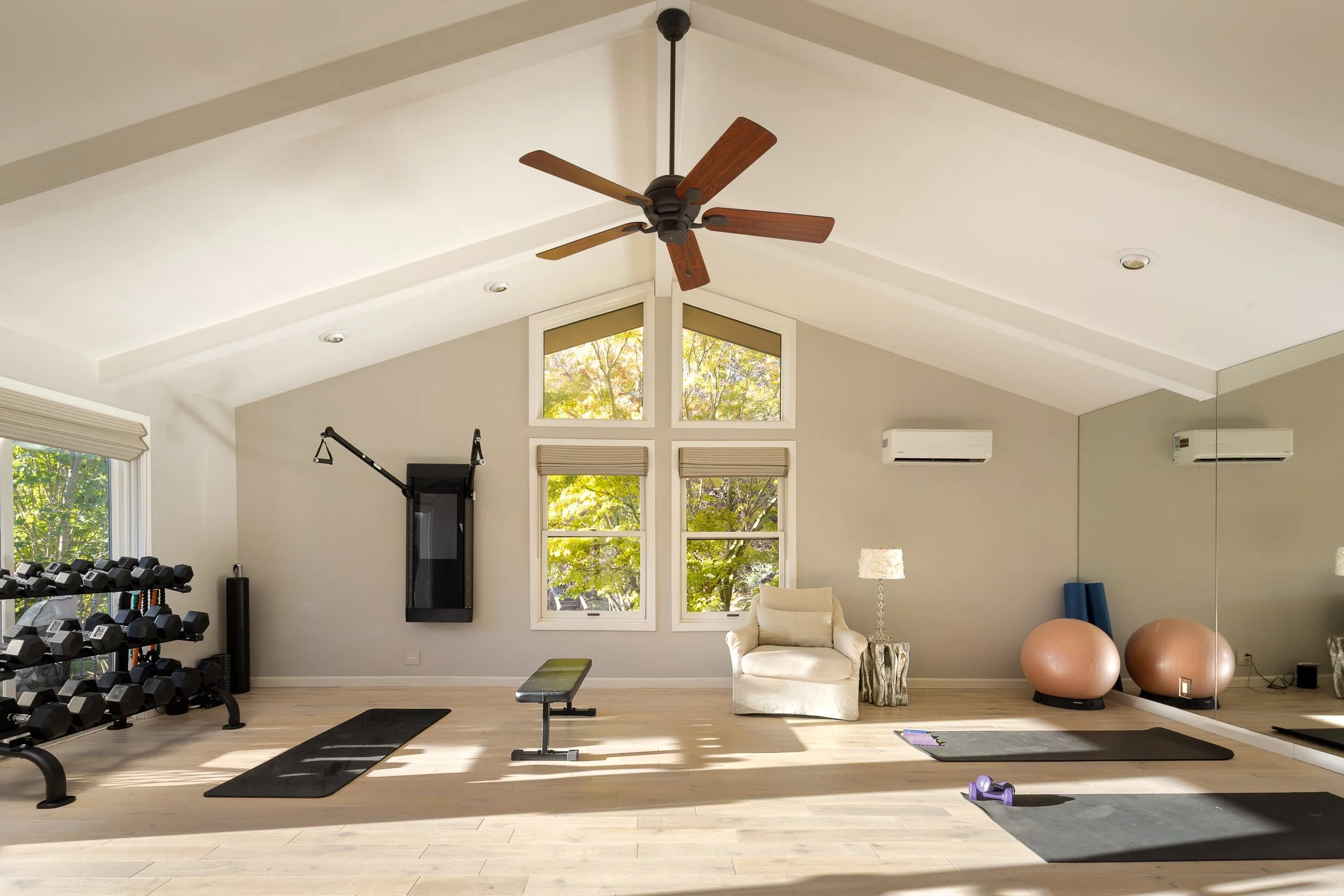 Home gym with dumbbells, exercise bench, yoga mats, exercise balls, and a comfortable chair by the windows with natural light.