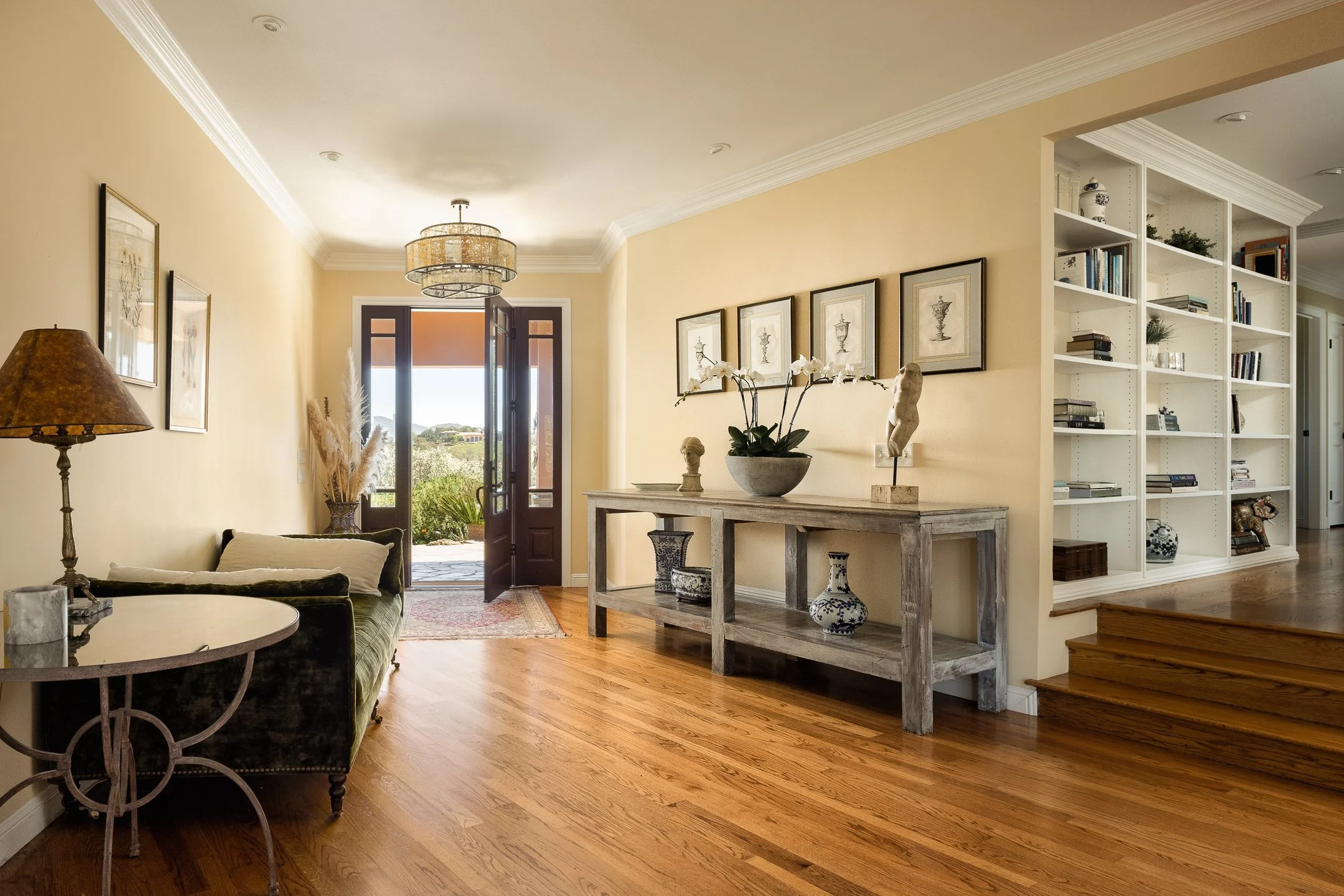 A living room with yellow walls, hardwood floors, a dark sofa, a side table, artwork on the wall, a ceiling light fixture, a doorway with a view outside, and shelves with books and decorative items.