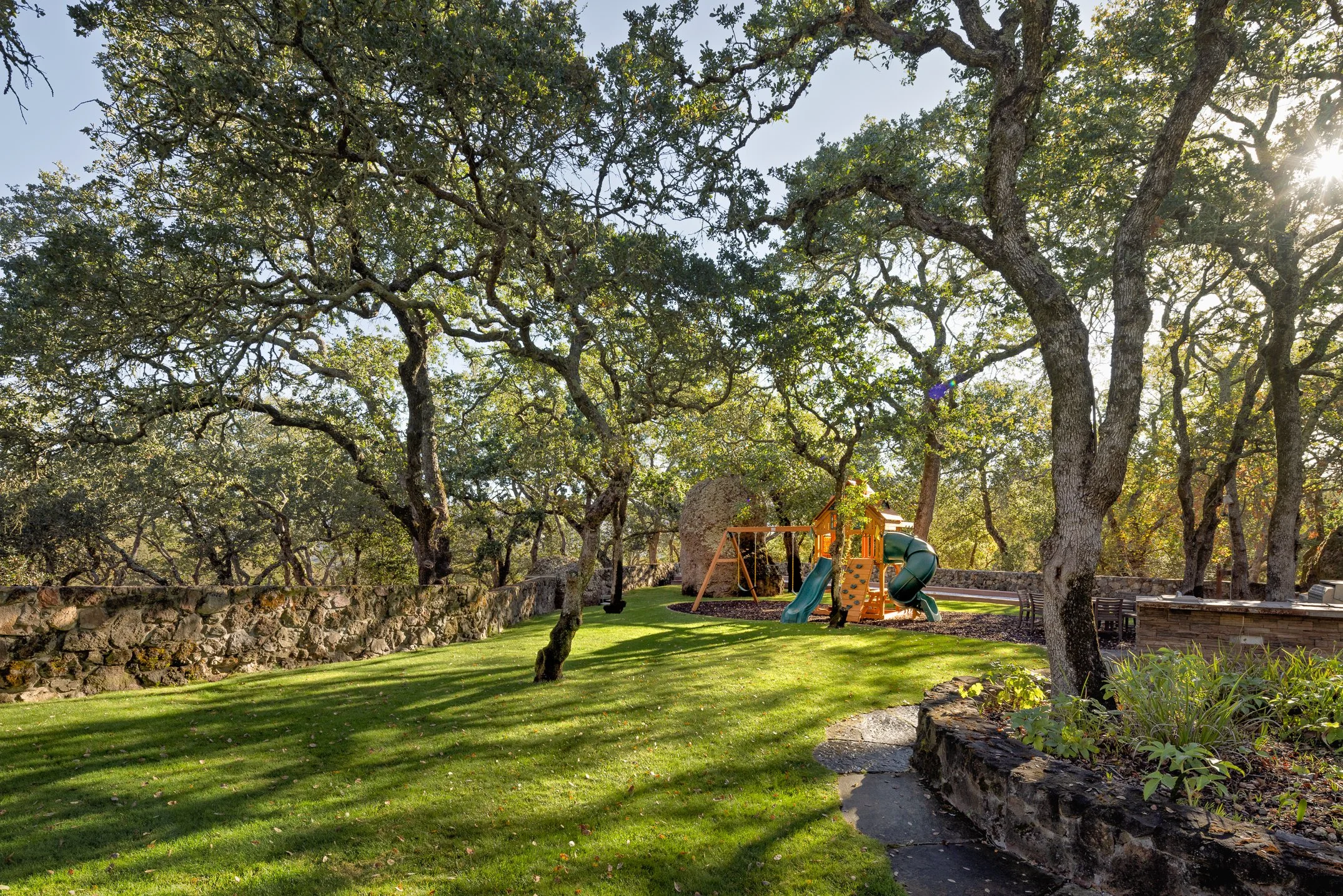 A backyard with tall, leafy trees providing shade. There is a small playground with a slide and swings, a stone wall, a bench, and a rock garden. The sun is shining through the trees.