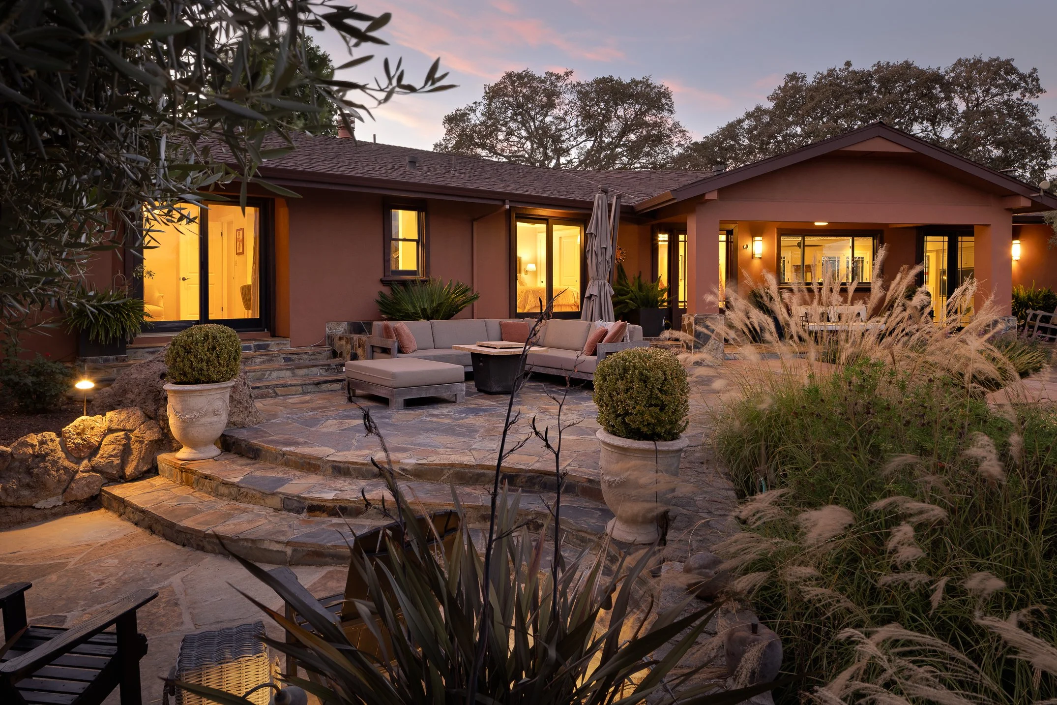 Exterior view of a house with large glass doors and windows illuminated from inside, surrounded by a landscaped backyard with potted plants, patio furniture, and decorative grasses at dusk.