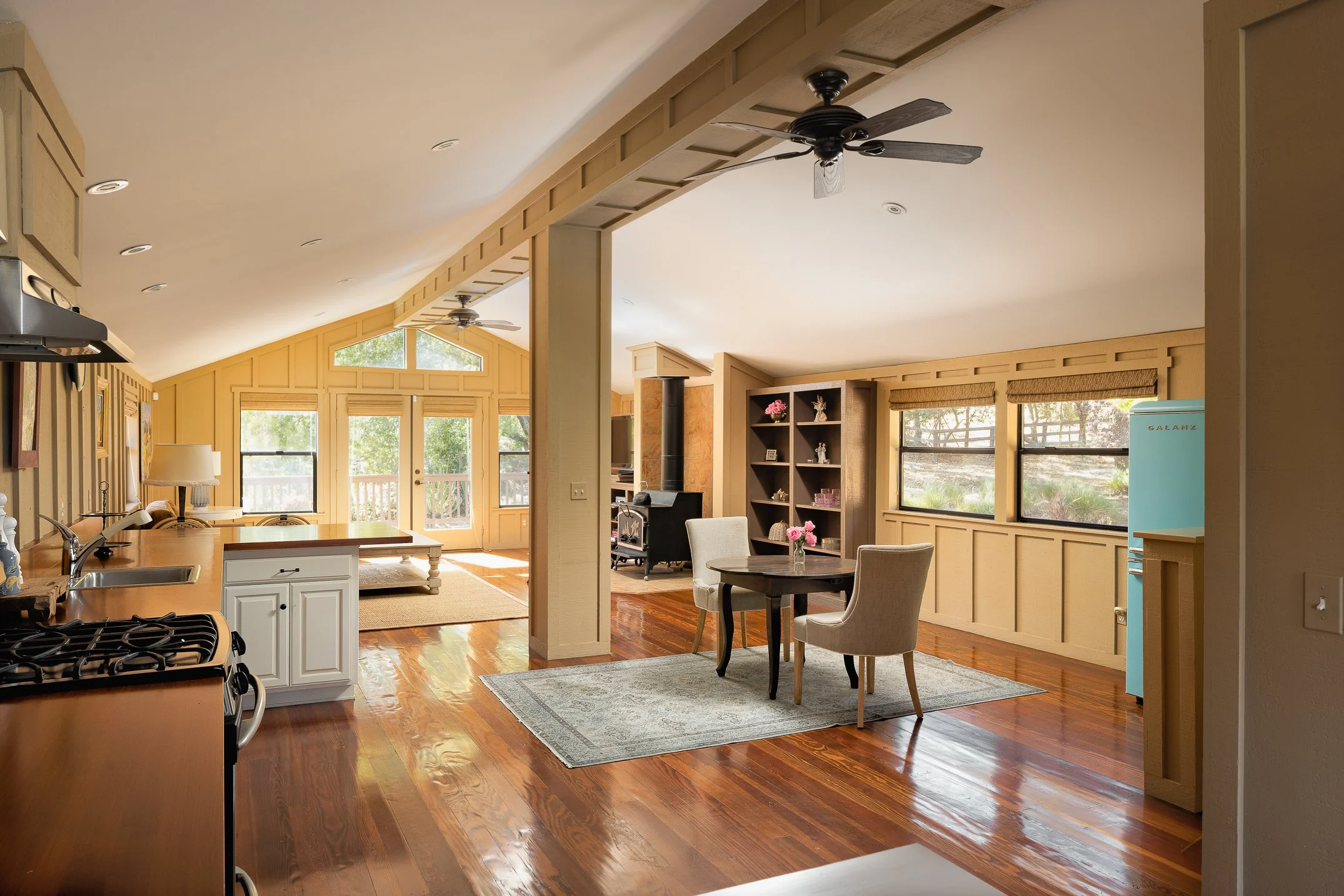 Interior view of a spacious living area with large windows, wooden flooring, beige walls, a kitchen on the left, and a dining area with a round table and four chairs, some decorated with flowers, and ceiling fans.