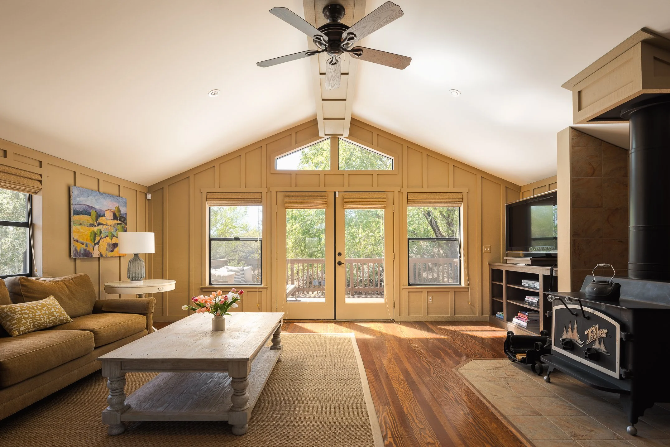 Living room with beige walls, large windows, a ceiling fan, a beige sofa, a white coffee table with a flower arrangement, and a TV with a fireplace.