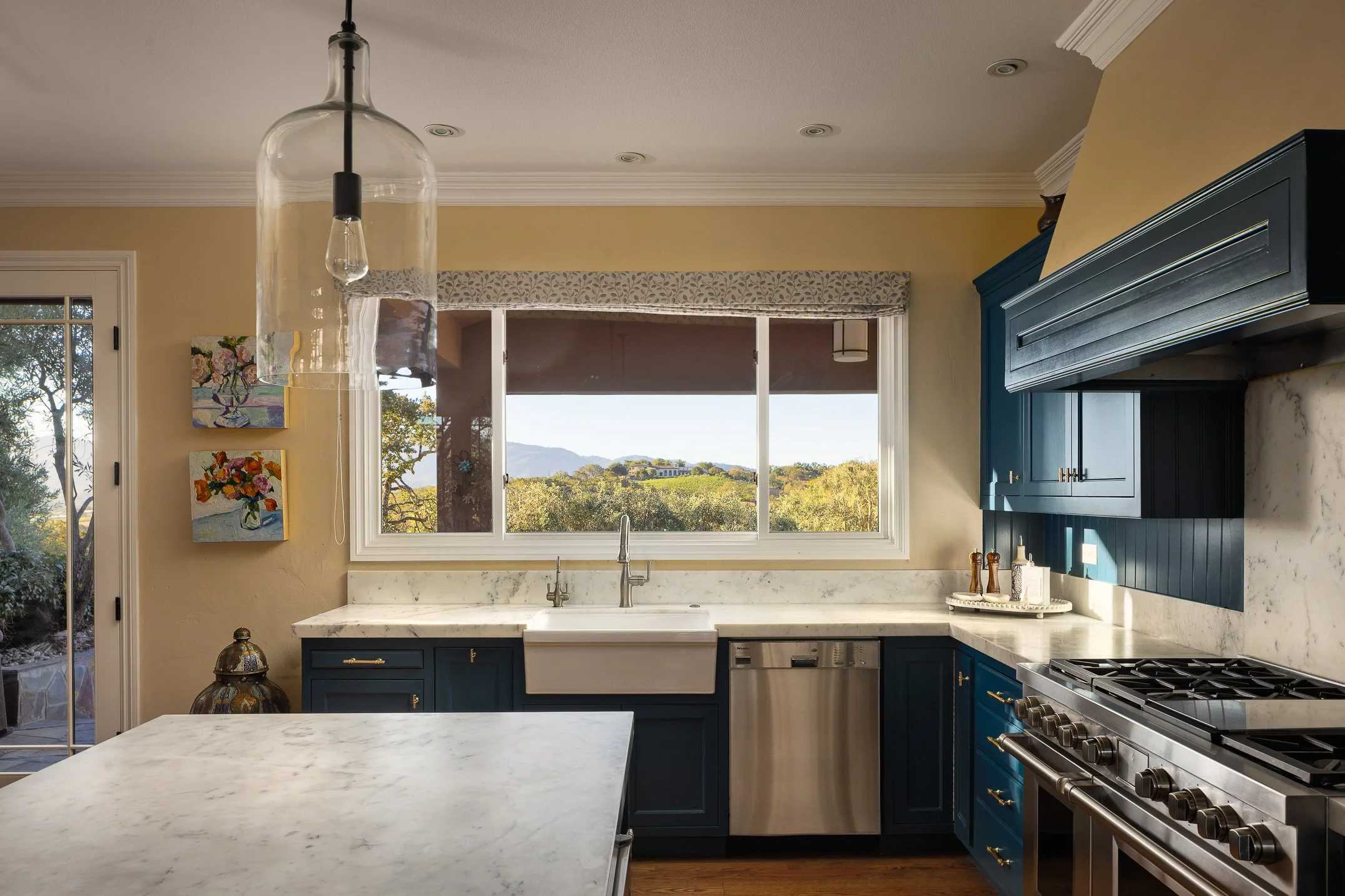 A modern kitchen with white marble countertops, dark blue cabinets, a large window showing a scenic view of Mt. George, and artwork on the yellow walls.