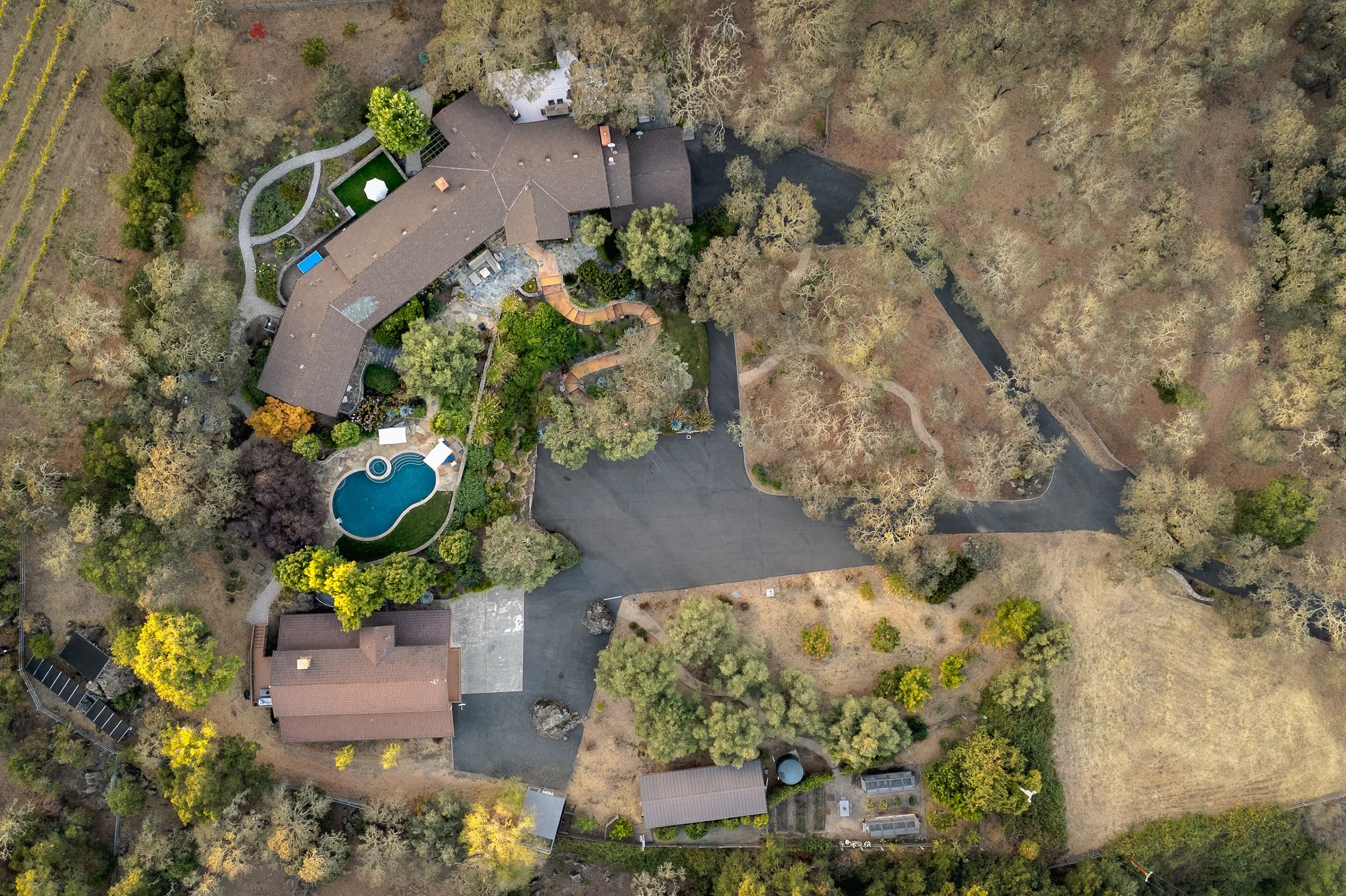 Aerial view of a residential house surrounded by trees, with a swimming pool and garden area, and with a road and open land nearby.
