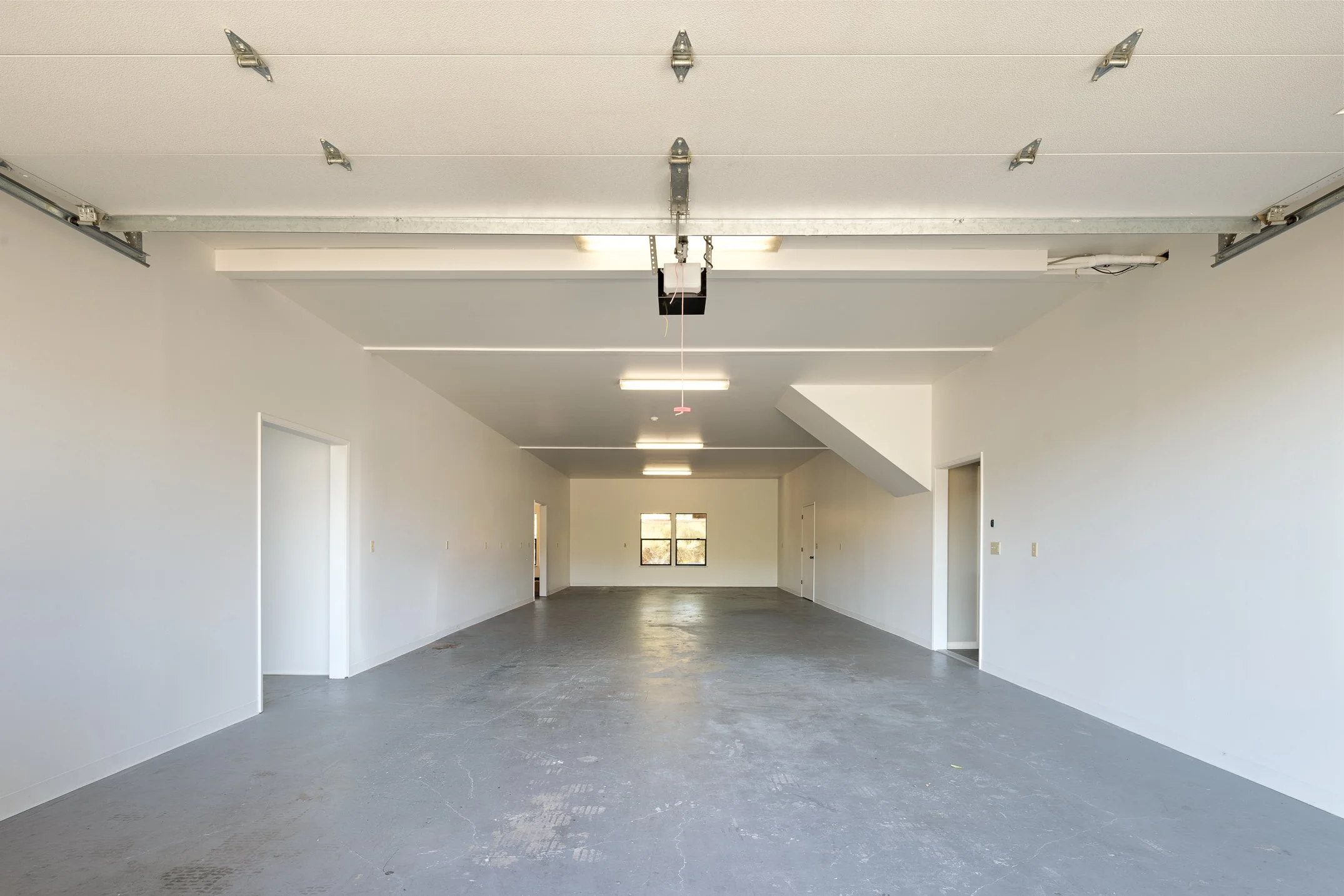 Empty garage with white walls, gray concrete floor, ceiling lights, and open garage door.
