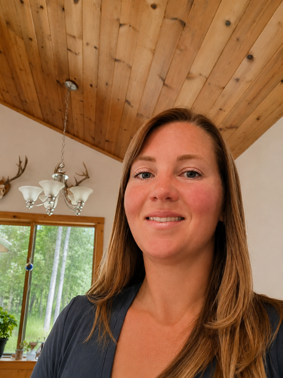 Close-up of a smiling woman with long brown hair inside a room with wooden ceiling and a large window showing greenery outside.