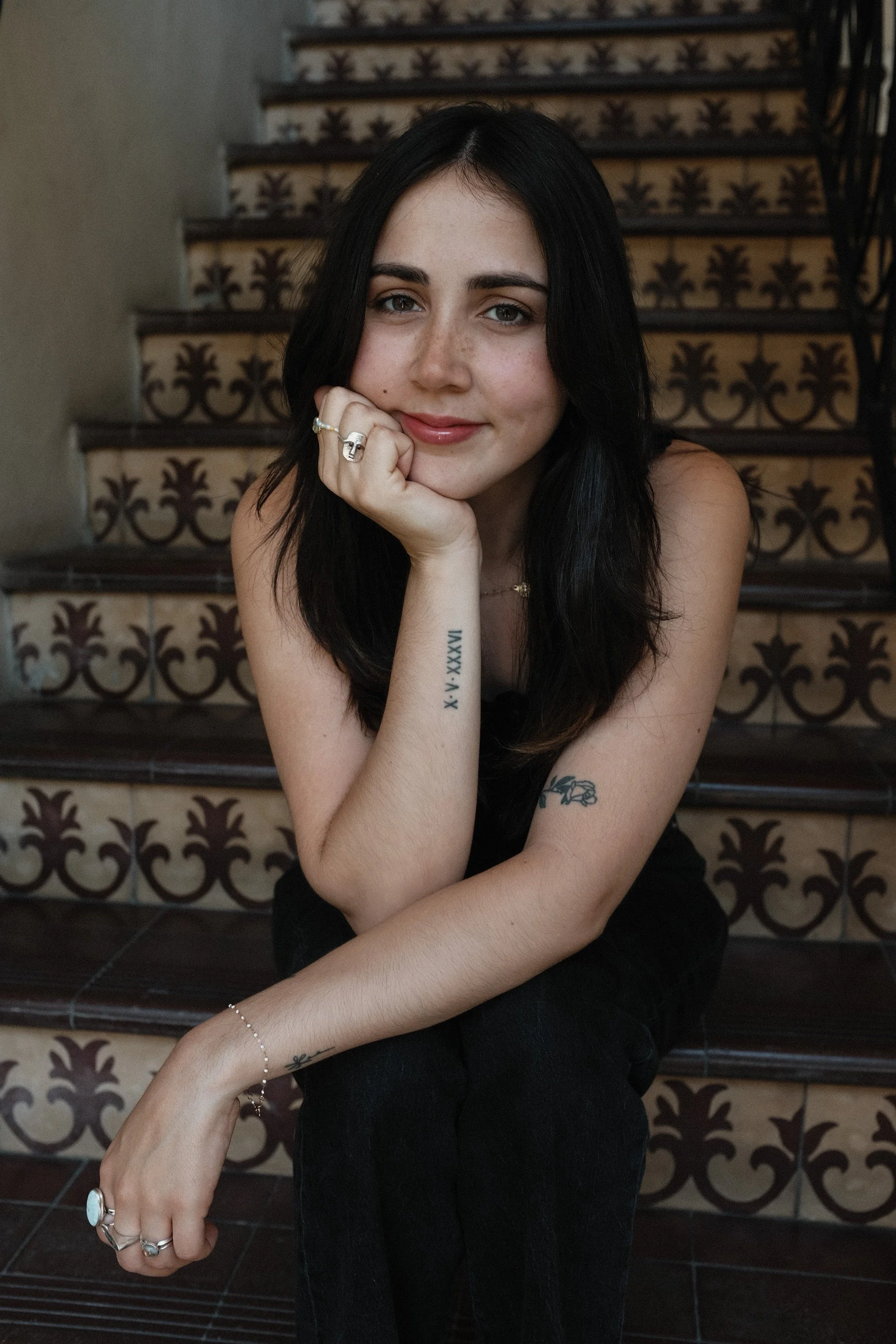 A young woman with black hair, fair skin, and light makeup, sitting on tiled stairs with ornate ironwork on the sides, resting her chin on her hand, wearing jewelry including rings, a bracelet, and a necklace.