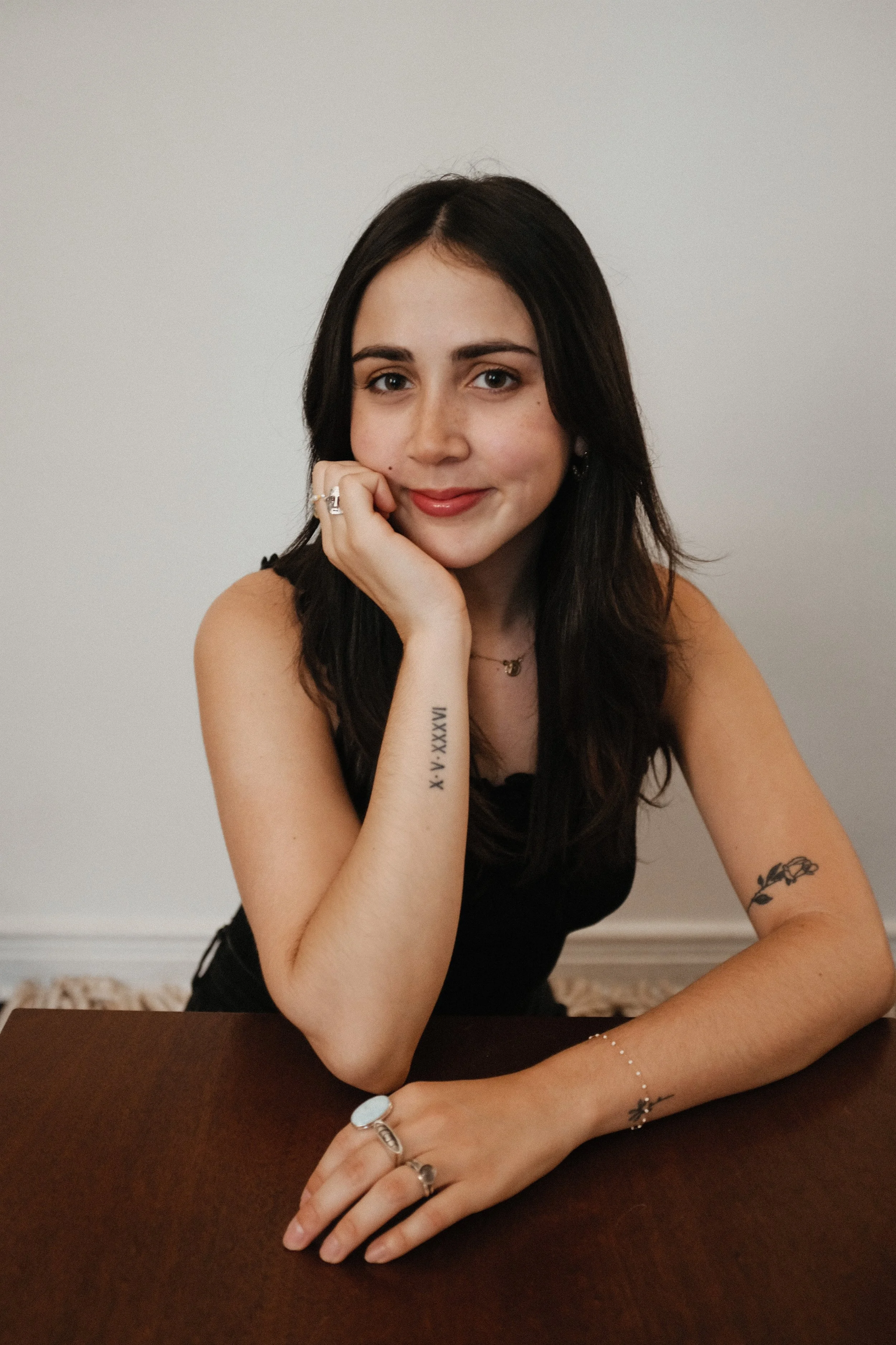 A woman with dark hair, light skin, and tattoos resting her chin on her hand, smiling softly at the camera, sitting at a table with a plain white wall background.
